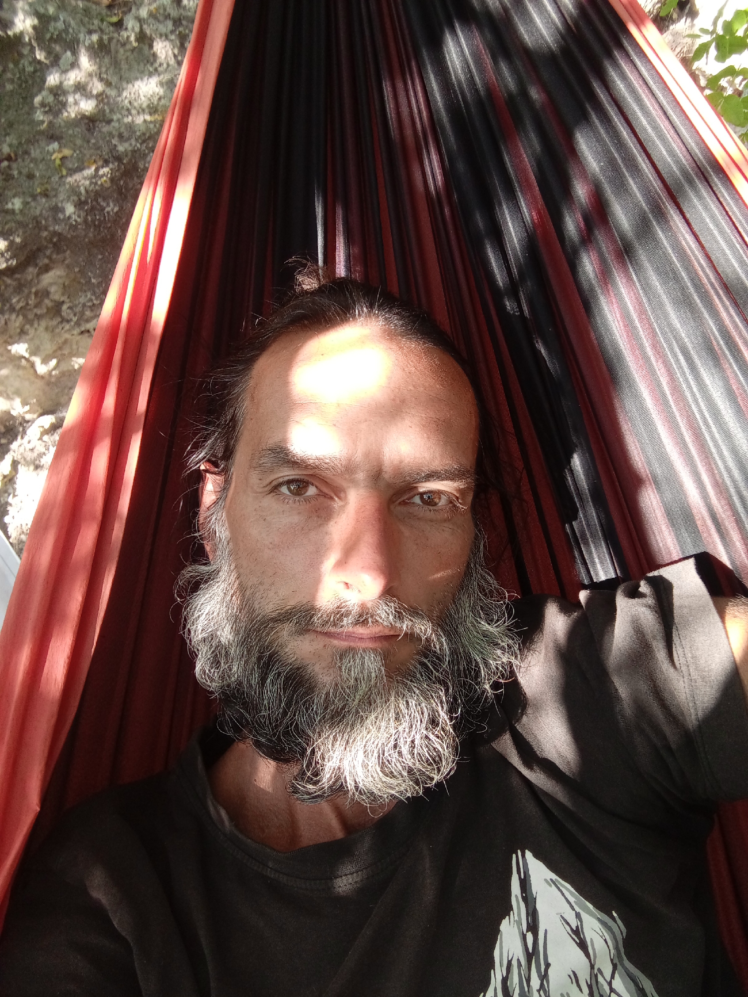 A close-up, overhead shot shows a man with long, dark hair tied in a bun, a full grey beard, and a beard on his chin. He is lying in a hammock with red and black straps. The man is wearing a dark t-shirt with a white graphic of a mountain range. Sunlight streams through the hammock straps, casting shadows on his face and body. The background shows a rocky surface and foliage, possibly suggesting an outdoor location in Балаклавский округ, Ukraine. The man is looking directly at the camera with his eyes open and a neutral expression.