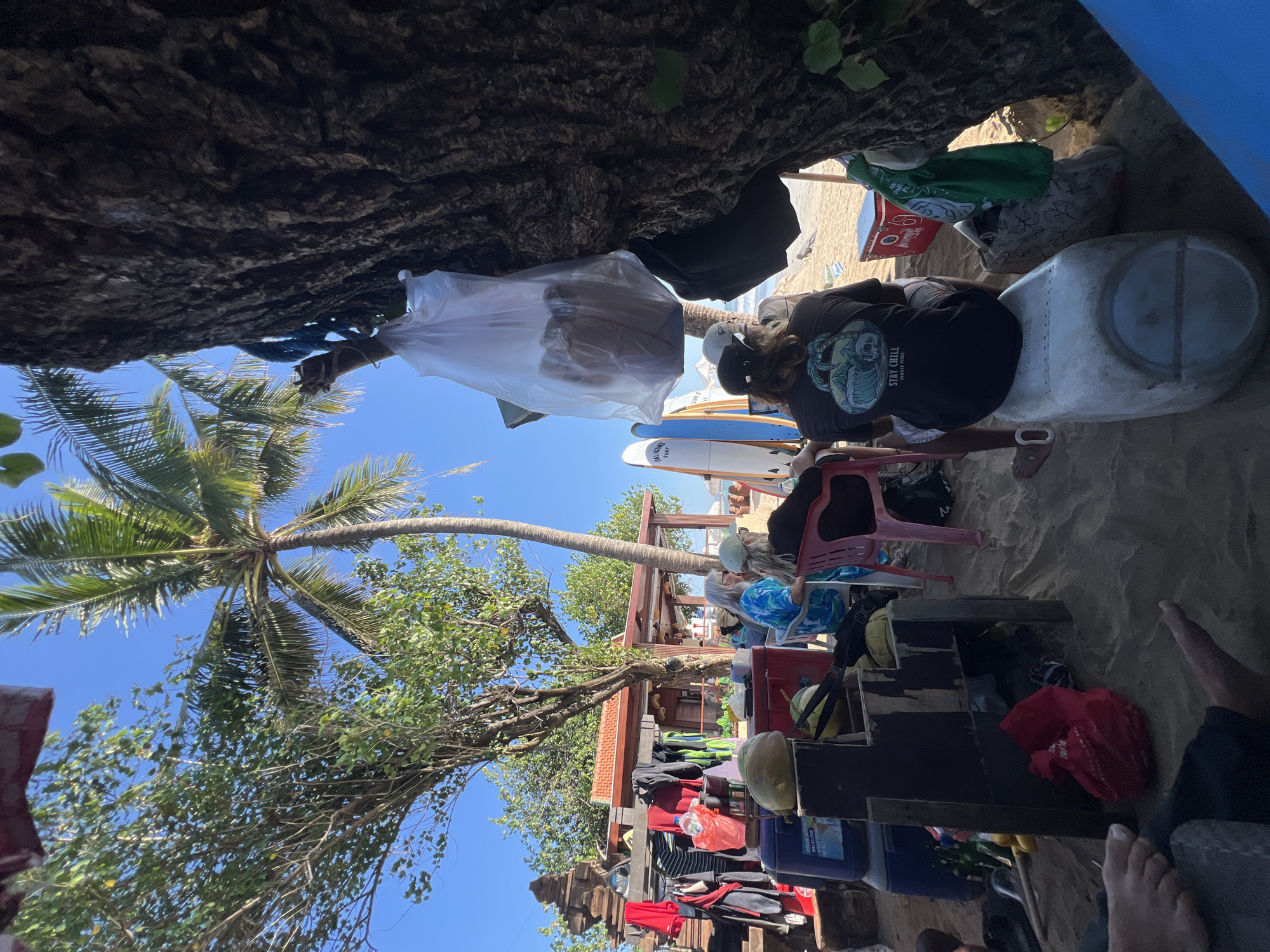 In Kuta, Indonesia, a group of individuals is gathered on a sandy beach near a small wooden structure adorned with a red tiled roof. Several surfboards stand upright, leaning against a tree trunk. A person wearing a black t-shirt with a graphic design of a skull and waves sits on a white plastic container. Other individuals, some with long hair, are seated on chairs. Various items are scattered around, including a large blue cooler, a red cooler, bags, clothing hanging on a rack, and coconuts. A white plastic bag filled with items is suspended from a tree branch. The scene suggests a relaxed atmosphere, possibly a break from surfing or a casual beach gathering.
