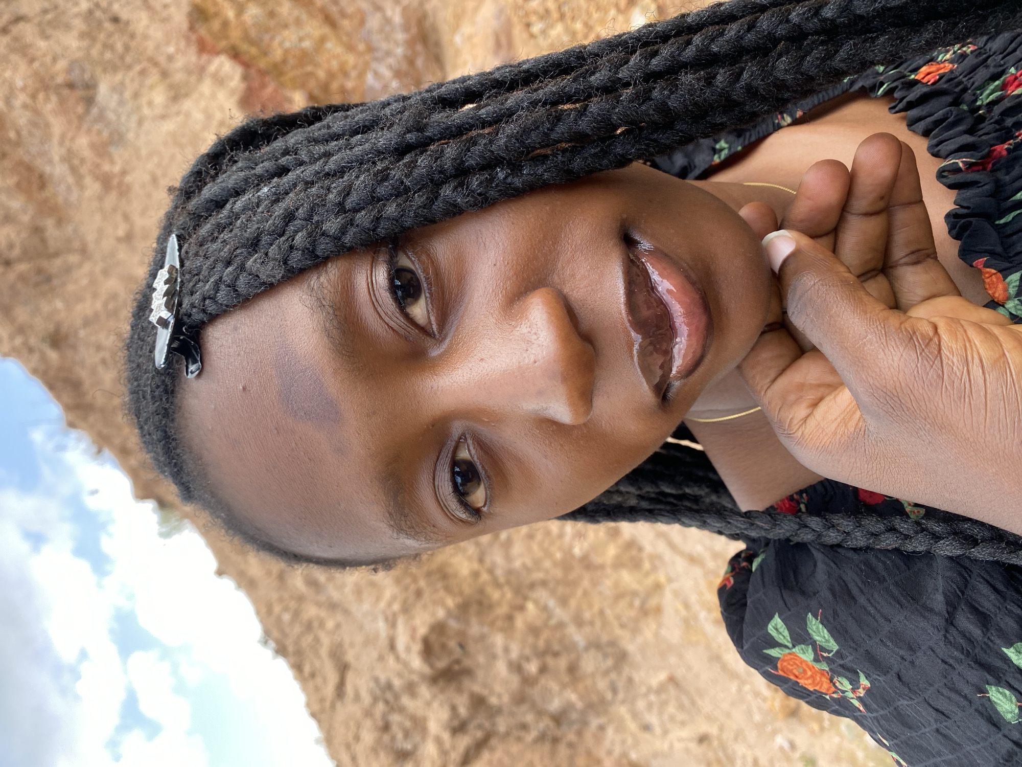 A close-up, outdoor, selfie-style portrait shows a young Black woman with dark braided hair, wearing a black dress with a floral pattern. She is outdoors with a sandy-colored cliff face and a cloudy sky in the background. The woman is holding her chin with her left hand. A silver barrette with a decorative pattern is visible in her hair. The overall lighting suggests it is daytime. The scene appears to be in a natural, possibly rural, environment.
