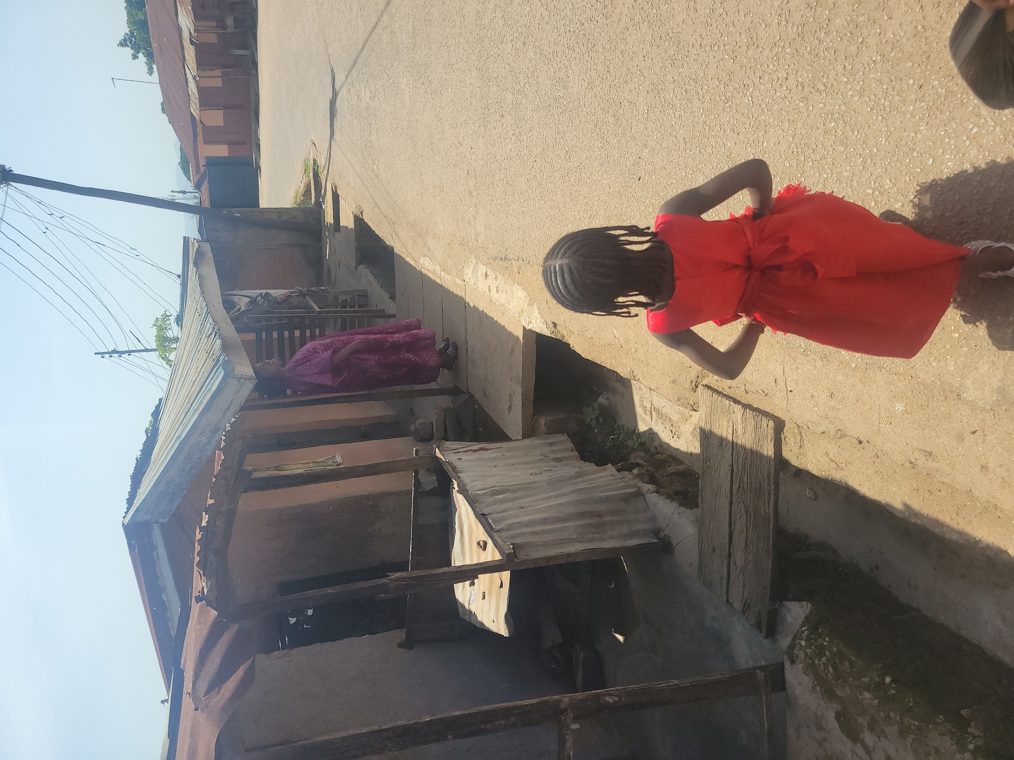 The scene is an outdoor shot taken in Jos, Nigeria. A young girl in a red dress stands on a sunlit paved road. Her back is to the camera, and her arms are slightly bent at the elbows with her hands resting on her hips. She has dark hair styled in braids. Next to the road are dilapidated buildings with rusty corrugated metal roofs. An adult female in a purple gown stands beside one of the structures. Utility poles with electrical wires are visible near the building on the left side of the image. The environment suggests an impoverished neighborhood.