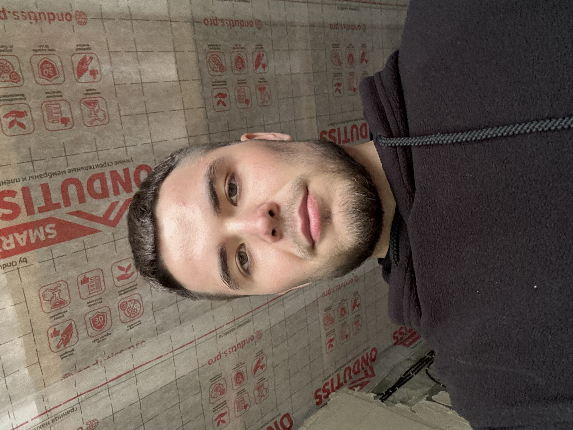 This is a portrait-oriented, slightly tilted, eye-level photograph of a young man with short dark hair and a beard. He is wearing a dark hooded sweatshirt. He is looking towards the camera with a neutral expression. The background consists of a translucent material printed with a grid pattern and various red icons and text. The text is in Russian and includes "ONDUTISS," "@ondutiss.pro," and other phrases like "умные строительные мембраны и пленки" (smart building membranes and films) and "граница нахлеста" (overlap line). The icons depict symbols such as a flask, a thumbs-up, a hand with a leaf, a document, a clock with "30" inside, and a pizza slice. The overall impression is that the man is standing in front of or leaning against a roll of construction material. The lighting appears to be artificial. There are no clear indicators of time of day or weather. The location context is Perm, Russia.