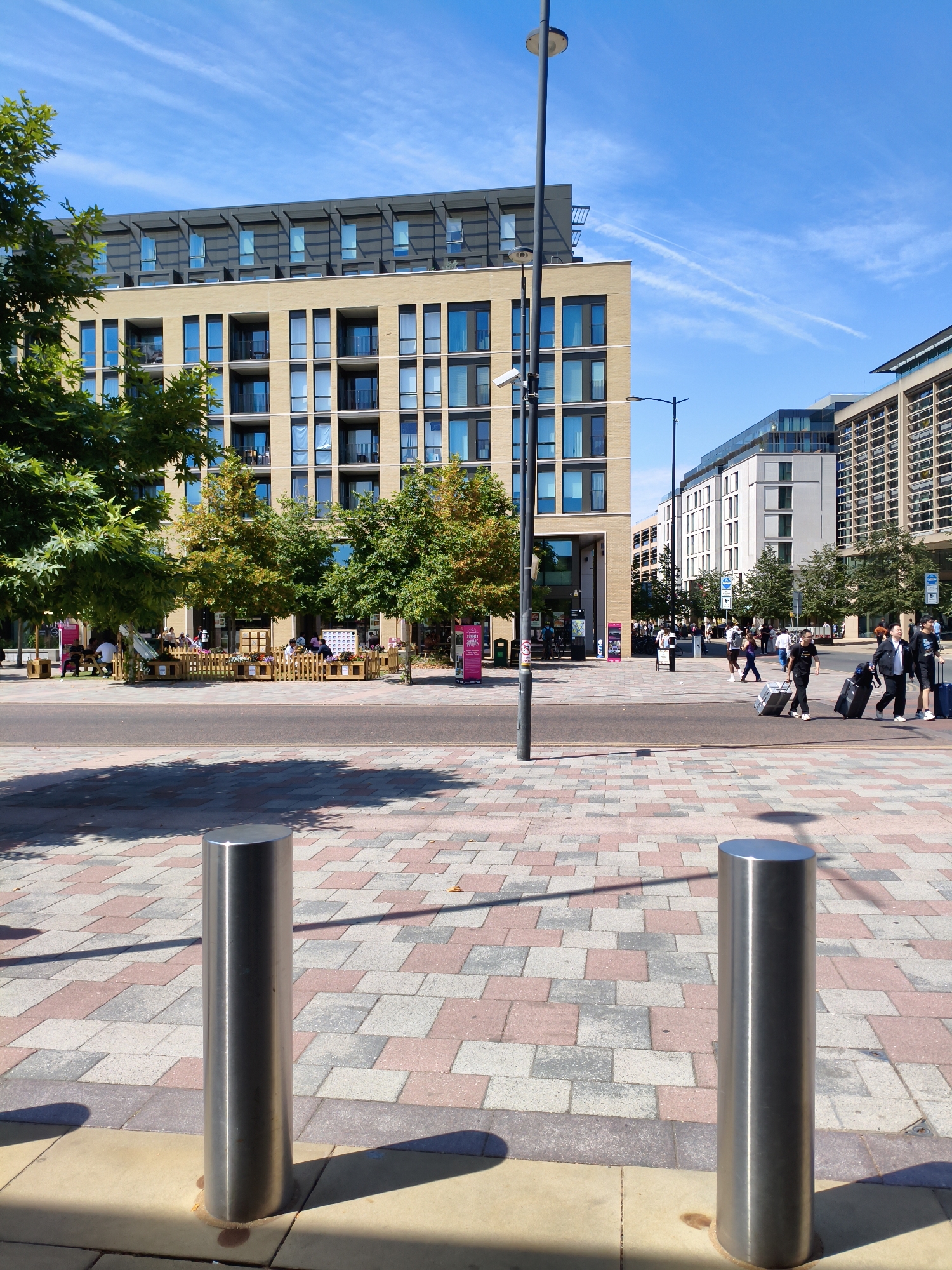 This is a medium shot, taken from a low angle, of a street scene in Cambridge, United Kingdom. The scene is dominated by a modern, multi-story building with a beige facade and blue-tinted windows, stretching across the background. In the foreground, a paved area with a checkerboard pattern of pink, grey, and beige tiles is visible. Two polished stainless steel bollards stand upright in the foreground, casting long shadows.
To the left of the frame, a large green tree with dense foliage partially obscures the building. In the middle ground, several people are seated at tables outdoors, separated by a low wooden fence. Beyond them, more trees with yellowing leaves add color to the scene.
Further into the background, other modern buildings with glass facades line the street. A lamppost with a CCTV camera attached stands prominently in the mid-left, and another lamppost is visible further down the street. Several people are walking along the pavement, some of whom are pulling luggage, suggesting they might be travelers. The sky is a clear, bright blue with a few wispy white clouds. The overall impression is of a sunny day in a contemporary urban environment.