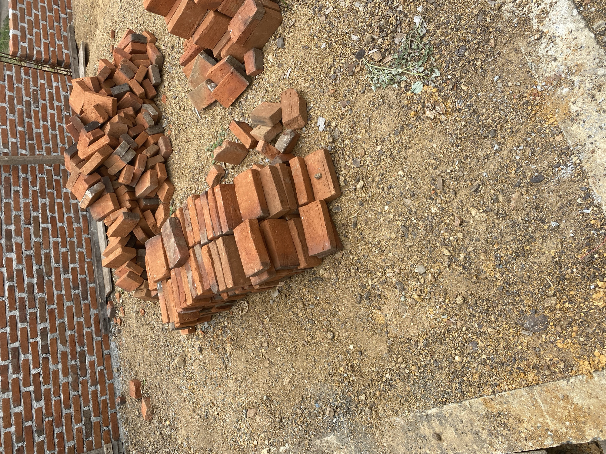 The image depicts a construction site in an unknown location. A brick wall is partially constructed in the background, with loose bricks and debris scattered on the sandy ground in the foreground. Several piles of red bricks are visible, suggesting they are either waiting to be used or have been discarded. The ground is covered with sand, gravel, and small stones, indicating an outdoor environment. There are no people or animals visible in the image. The scene suggests a typical construction process where building materials are organized and utilized.
