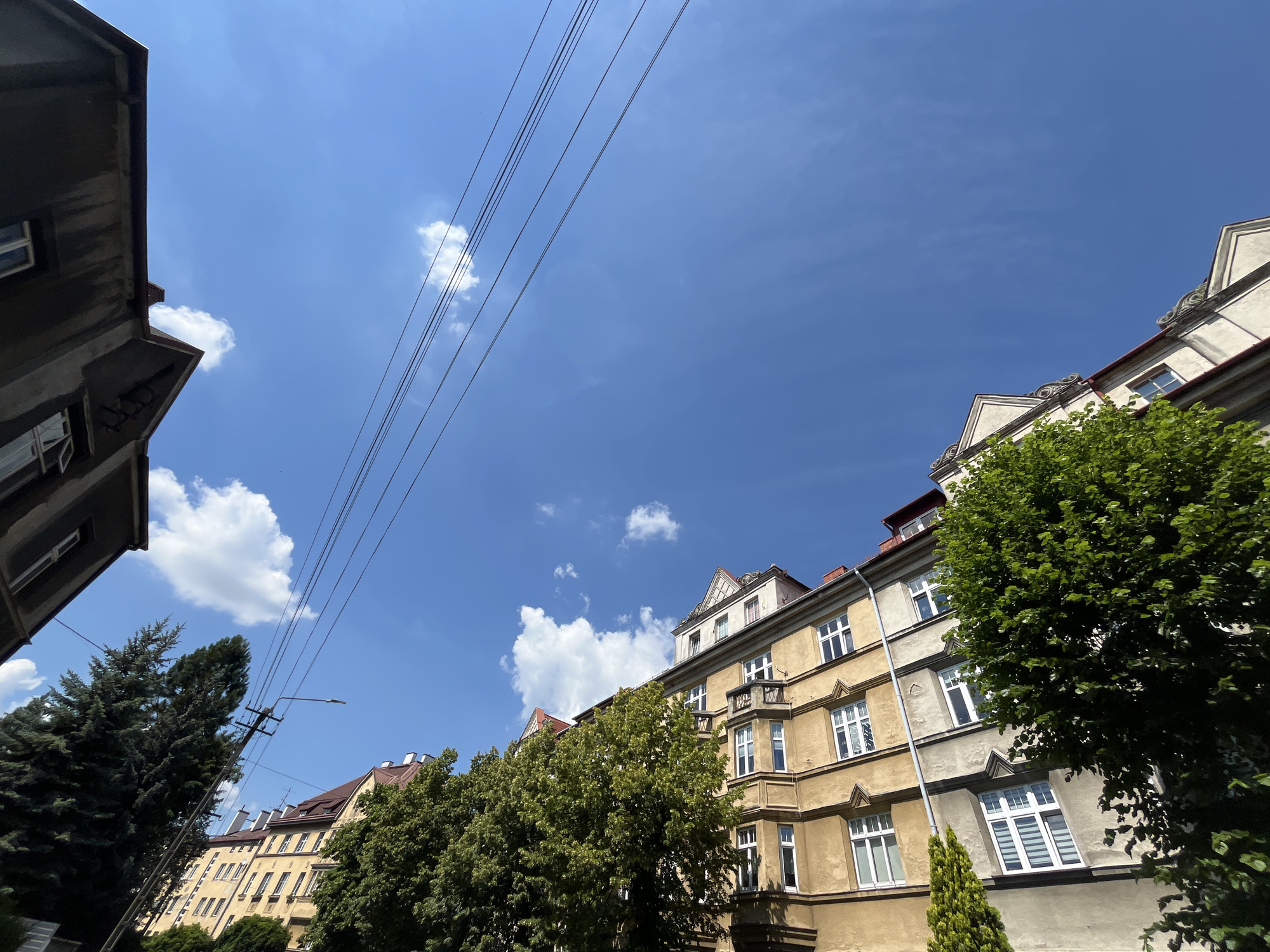 This media file shows an upward-angle shot of buildings and the sky. The scene is set outdoors, likely in an urban environment, with a clear blue sky dotted with fluffy white clouds. Several buildings are visible, with varying architectural styles and colors. One building in the foreground to the left appears to be darker, possibly made of concrete, with a window visible. To the right, a row of buildings with yellow and beige facades stretches into the distance. These buildings have multiple windows and some have balconies. Trees with lush green foliage are interspersed between the buildings and in the foreground, adding to the natural elements of the scene. Overhead, a series of power lines are strung across the sky, casting faint lines against the blue backdrop. The lighting suggests it is daytime, likely midday, given the brightness of the sky and the shadows. There is no discernible activity or event taking place, and no people are visible. The image evokes a sense of a normal day in a city, with a mix of man-made structures and natural greenery. No visible text is present in the image.