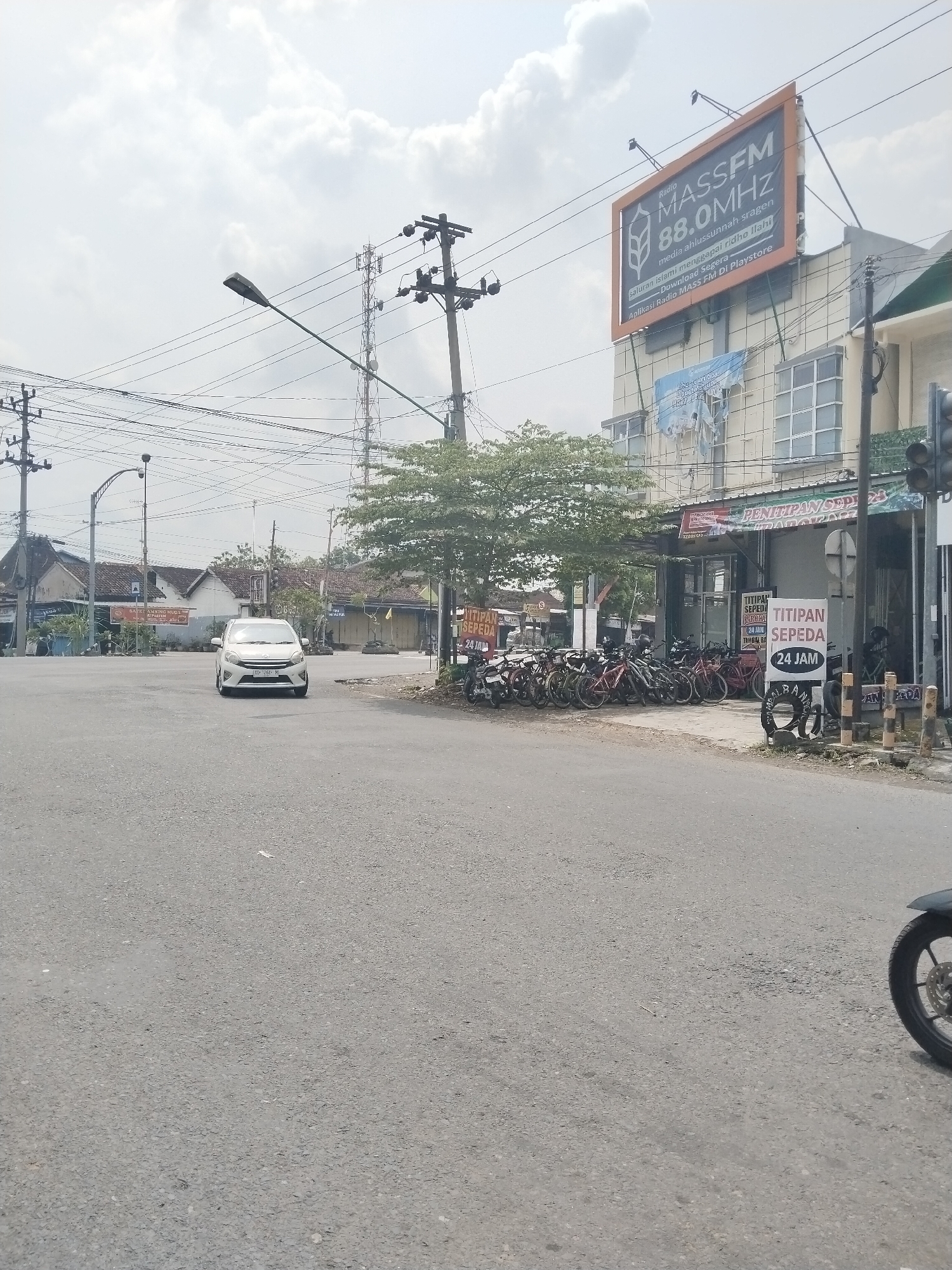 The image shows a street scene in an unidentified city in Unknown, Unknown. A white car is approaching the intersection. Overhead power lines crisscross the sky. On the right side of the road, a building features a large sign advertising "MASSFM 88.0MHz" radio. In front of the building, numerous bicycles are parked. Signs indicating "TITIPAN SEPEDA 24 JAM" (bicycle parking 24 hours) are visible. A motorcycle is partially visible in the lower right corner of the frame.