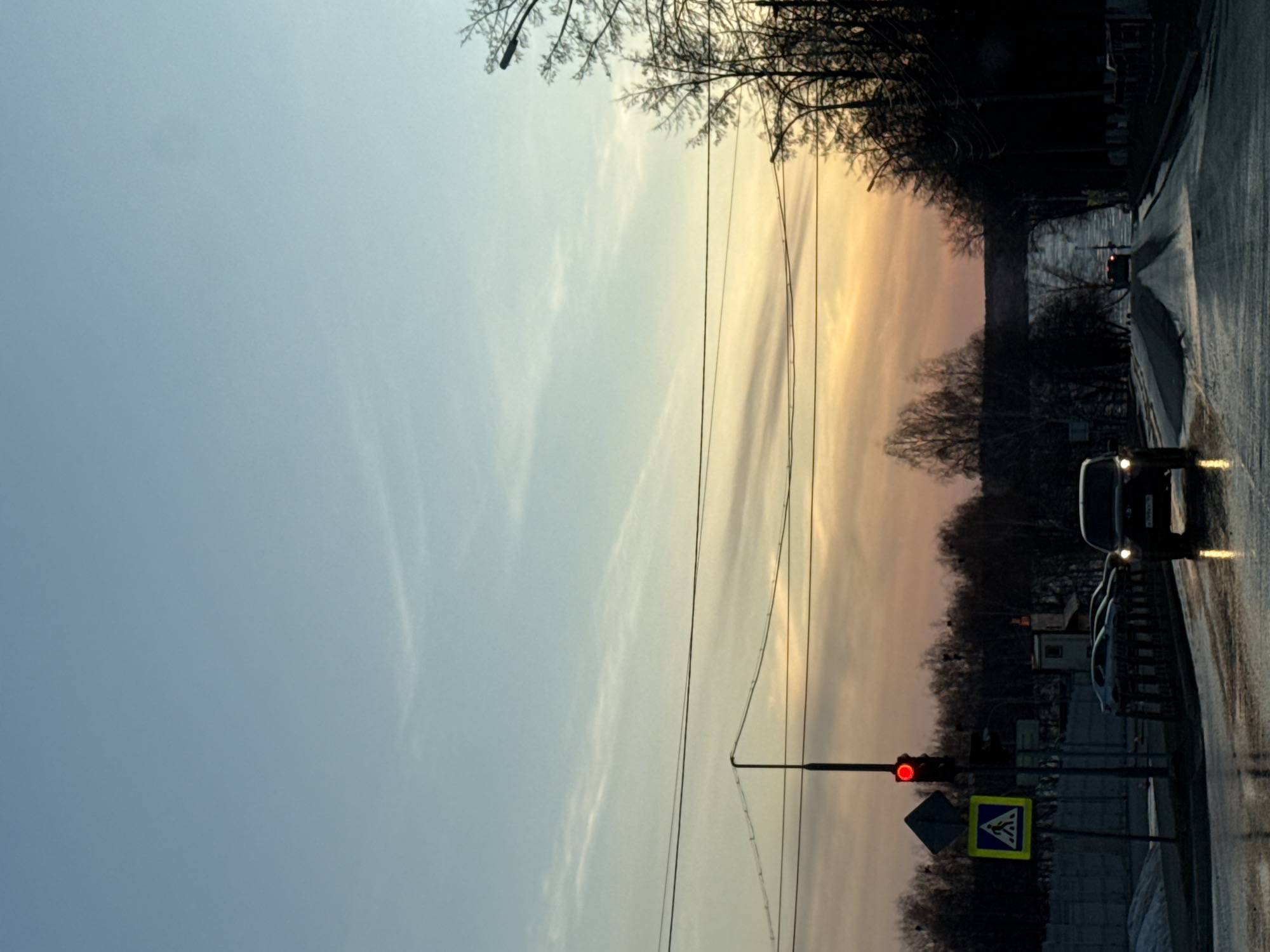 An eye-level shot depicts a road in Votkinsk, Russia, at dusk. The sky transitions from a light blue on the left to a gradient of orange and pink on the right horizon, visible above tree lines and buildings. Electrical wires stretch across the sky. A traffic light stands with a red light illuminated. A pedestrian crossing sign is visible. A car with headlights on is on the right side of the road. Other parked cars are also present along the road. The road is wet. Trees line the street.