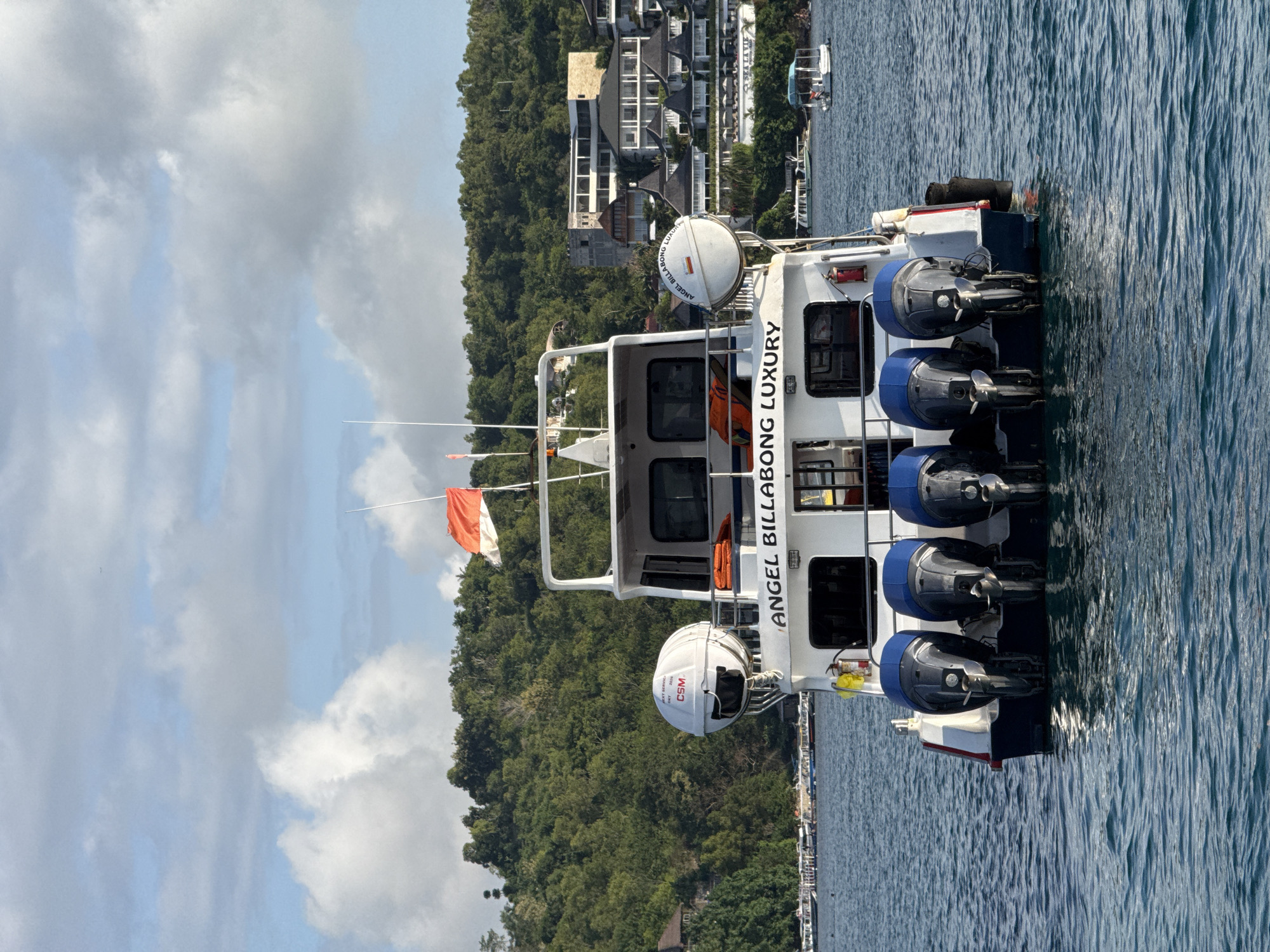 A white and blue speedboat, named "ANGEL BILLABONG LUXURY" as indicated by prominent text on its side, rests in calm, blue-green water. The vessel is equipped with at least five large, dark-colored outboard motors mounted at its stern, and two spherical white life rafts or bouys are positioned on its upper deck. One of these spheres displays "OXY," "CSM," and "SVCE" along with the boat's name in a circular pattern. An orange and white flag flies from a mast at the front of the boat's upper structure, and orange life jackets are visible through cabin windows. A yellow fire extinguisher is also mounted on the deck.
In the background, a steep, lush green hillside densely covered with trees rises from the water's edge. Modern buildings with distinctive roofs and architecture are nestled among the trees and along the shoreline, suggesting a resort or residential area. Another smaller boat can be seen further in the distance near the shore. The sky above is partly cloudy, with areas of bright blue peeking through the soft grey clouds. The scene depicts a bright daytime setting with calm conditions, characteristic of a tropical coastal environment like Kampung Toyapakeh, Indonesia.