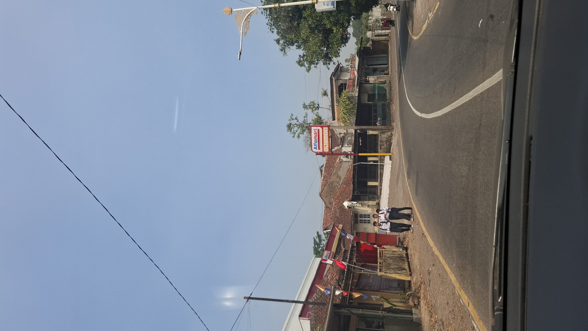 The image captures a street scene in Cilaku, Indonesia, likely on a sunny day. In the foreground, a dark car's dashboard is visible, suggesting the photo was taken from inside a vehicle. The road curves to the right, with a yellow line on the left edge and a white lane divider.
On the left side of the road, there are several buildings with traditional Indonesian architecture, featuring reddish-brown tiled roofs. Some buildings appear to be shops or small businesses, with one prominently displaying an "Alfamart" sign and promotional materials. In front of one building, there's a white fence and some colorful flags or banners. Three young people, likely students in school uniforms (white shirts and dark pants), are walking on the sidewalk.
The right side of the road features more buildings and lush green trees. A modern street lamp with an ornate design stands near the right edge of the frame. Further down the road, a motorcycle is parked or being ridden, and another building with a similar architectural style is visible. The sky is clear blue with a faint horizontal streak. The overall impression is a snapshot of daily life in a semi-urban or suburban area of Cilaku.