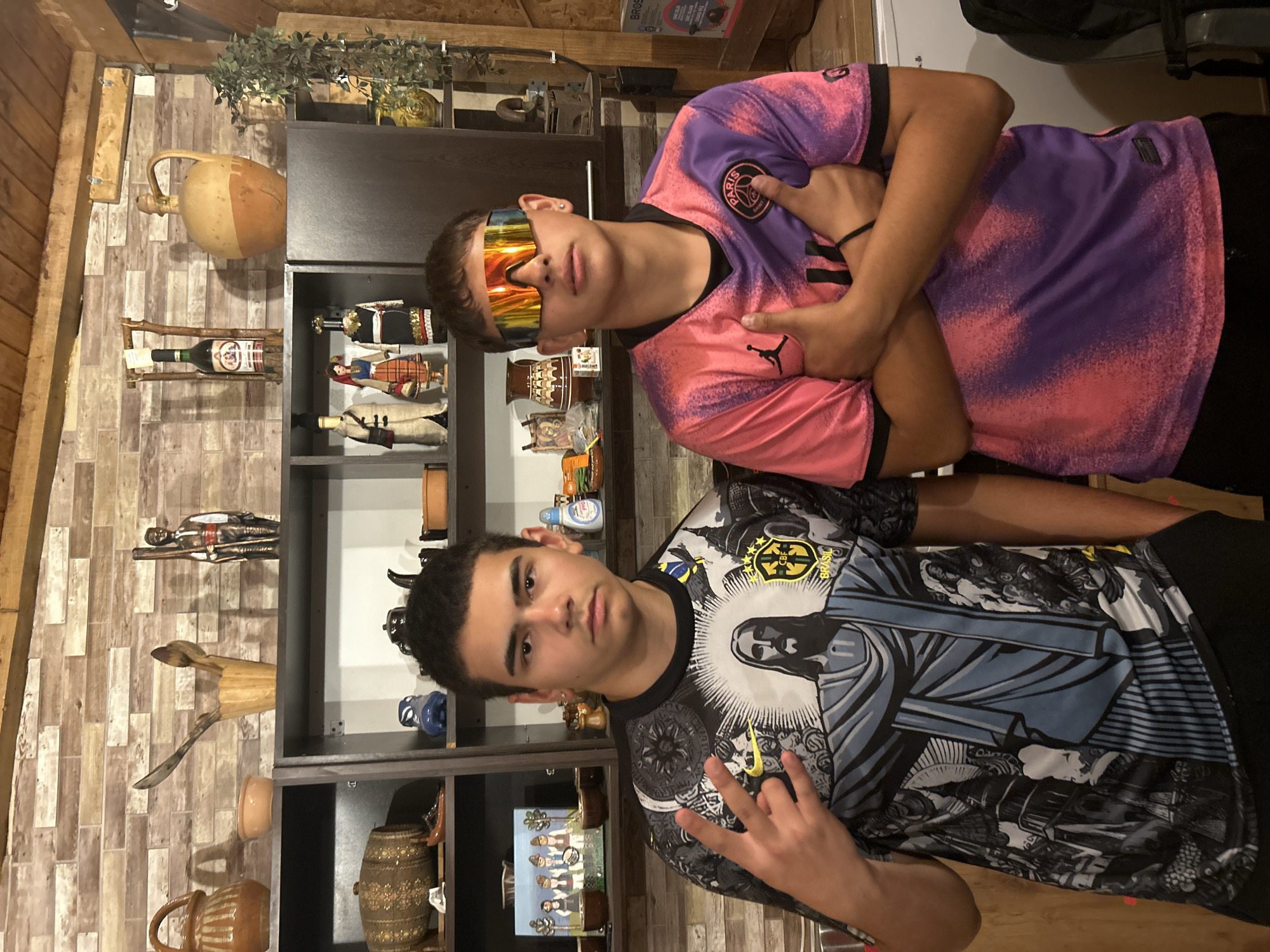 Two young males stand in front of a shelf displaying various decorative items. The male on the left wears a dark t-shirt with a graphic print of Jesus and cityscapes. He is making a peace sign with his right hand. The male on the right wears a pink and purple tie-dye t-shirt and large, mirrored sunglasses. He has his arms crossed. The shelf behind them contains pottery, dolls, bottles, and other assorted decorative objects. The background wall is a textured brick pattern.