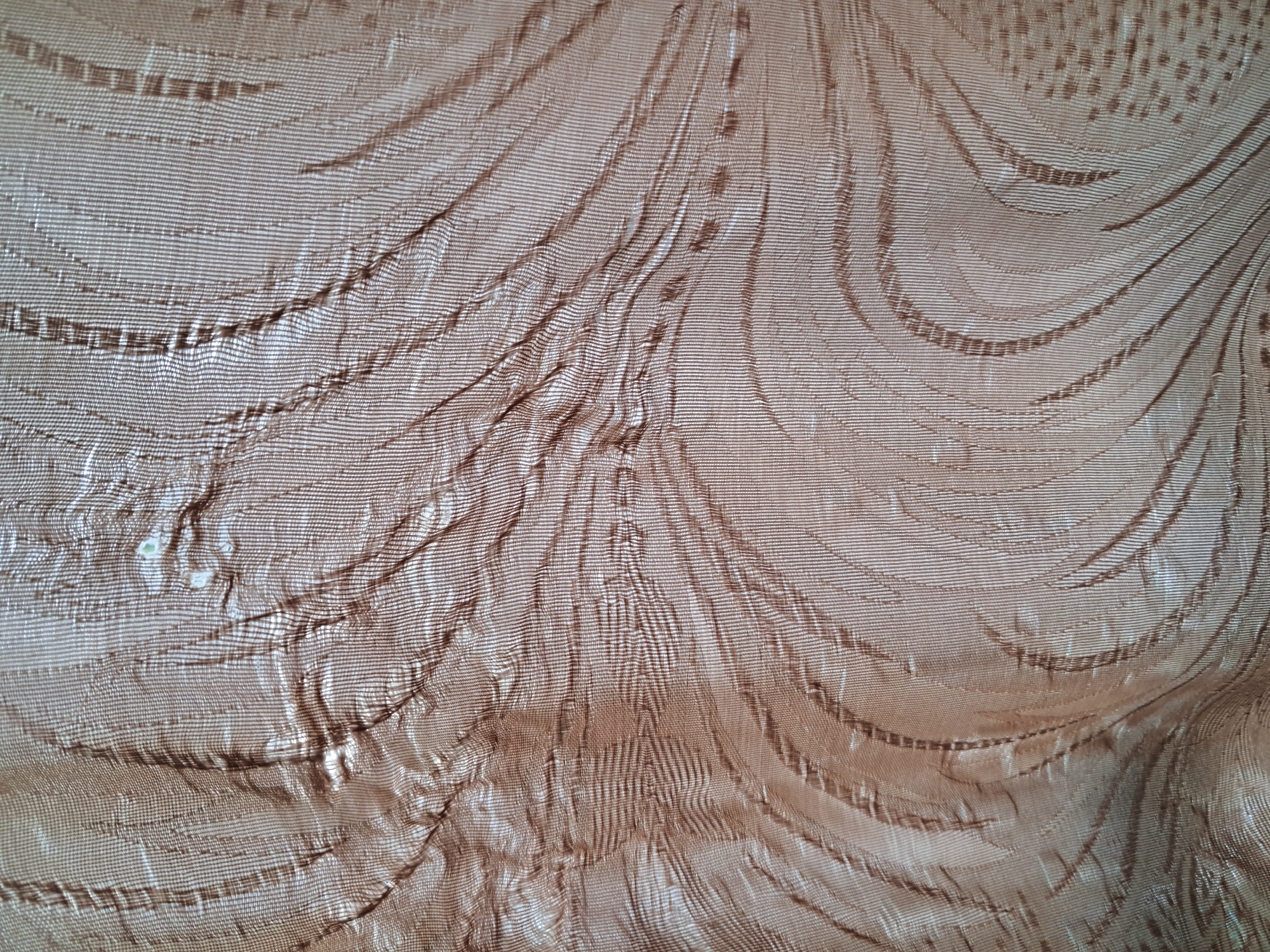 The image shows a close-up of a brown, textured fabric. The fabric appears to be sheer or semi-sheer, with a subtle sheen that reflects light in varying intensities, creating a dynamic play of highlights and shadows. The texture is characterized by vertical ribbing, which is then overlaid with a patterned design. This pattern consists of organic, flowing lines that resemble feathers or stylized waves, rendered in a slightly darker shade of brown or a different weave that creates depth. The fabric is creased and folded in several places, indicating it is likely a curtain or drapery. The overall impression is one of elegance and subtle luxury.
There is no information available to associate this image with the city of Bagish, Uzbekistan.