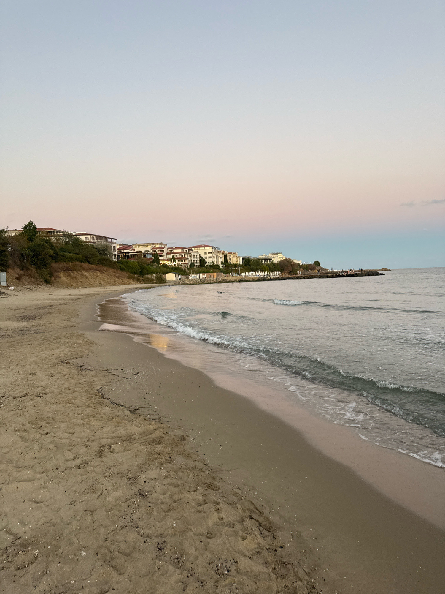 The image depicts a beach in Sveti Vlas, Bulgaria, with a gentle shoreline meeting the calm sea under a pastel-colored sky. The sand is a light tan, with scattered shells and debris. Along the upper edge of the beach, a grassy embankment slopes upwards, topped by a row of white buildings with terracotta roofs, indicative of a coastal resort. The sea is a muted gray-green, with small waves lapping at the shore. A man-made breakwater or pier extends into the sea on the right side of the frame, with a few figures visible along its length. The overall atmosphere is serene and tranquil, suggesting a quiet evening or early morning.