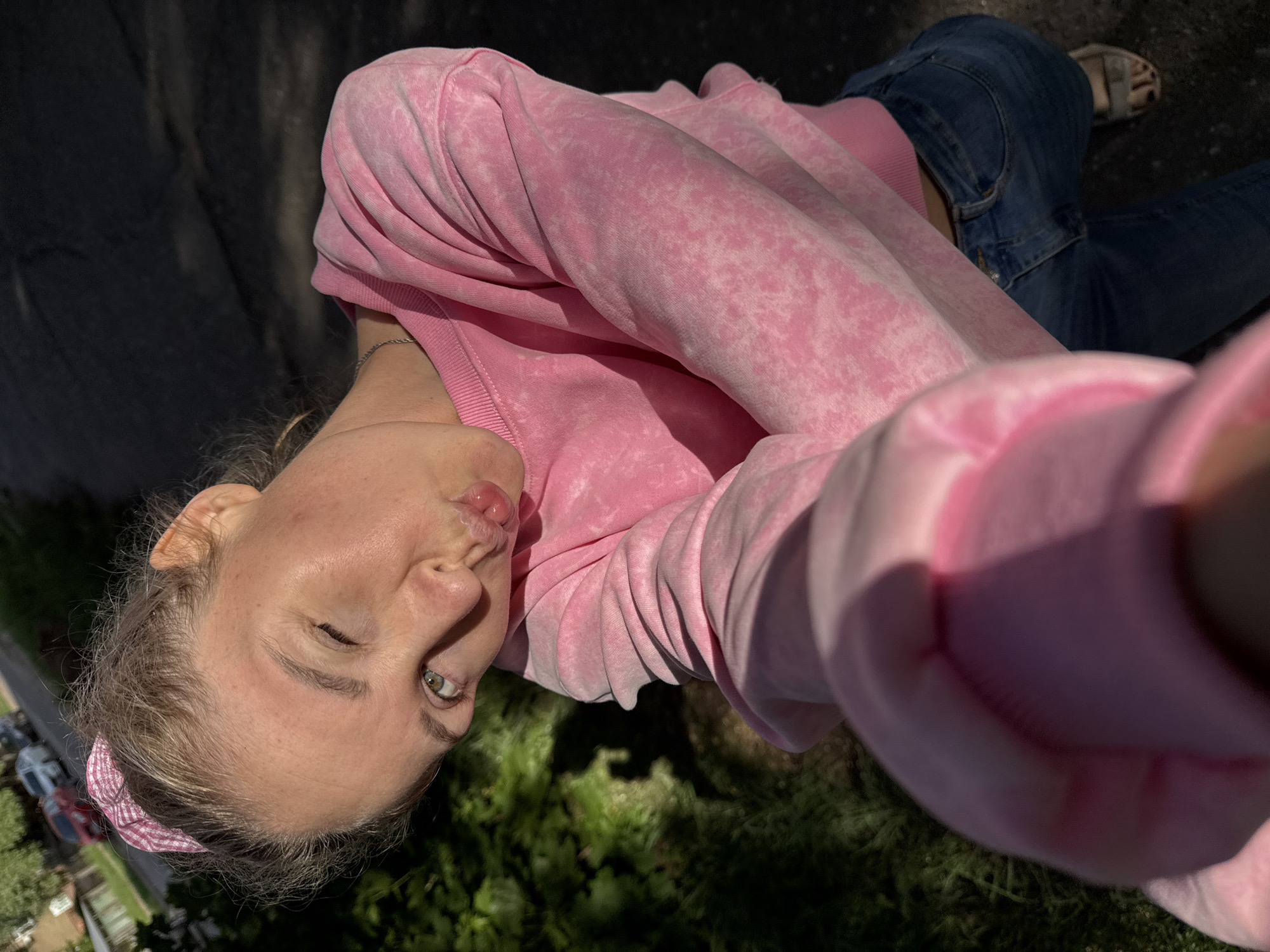 A selfie shows a young woman wearing a pink sweatshirt and blue jeans with a sandal on one foot. She is outdoors in Chaikovsky, Russia, as indicated by geolocation data. The background includes a street, grass, and some trees. Her head is tilted. She is making a duck-like expression with her lips. A pink and white checkered headband is visible.
