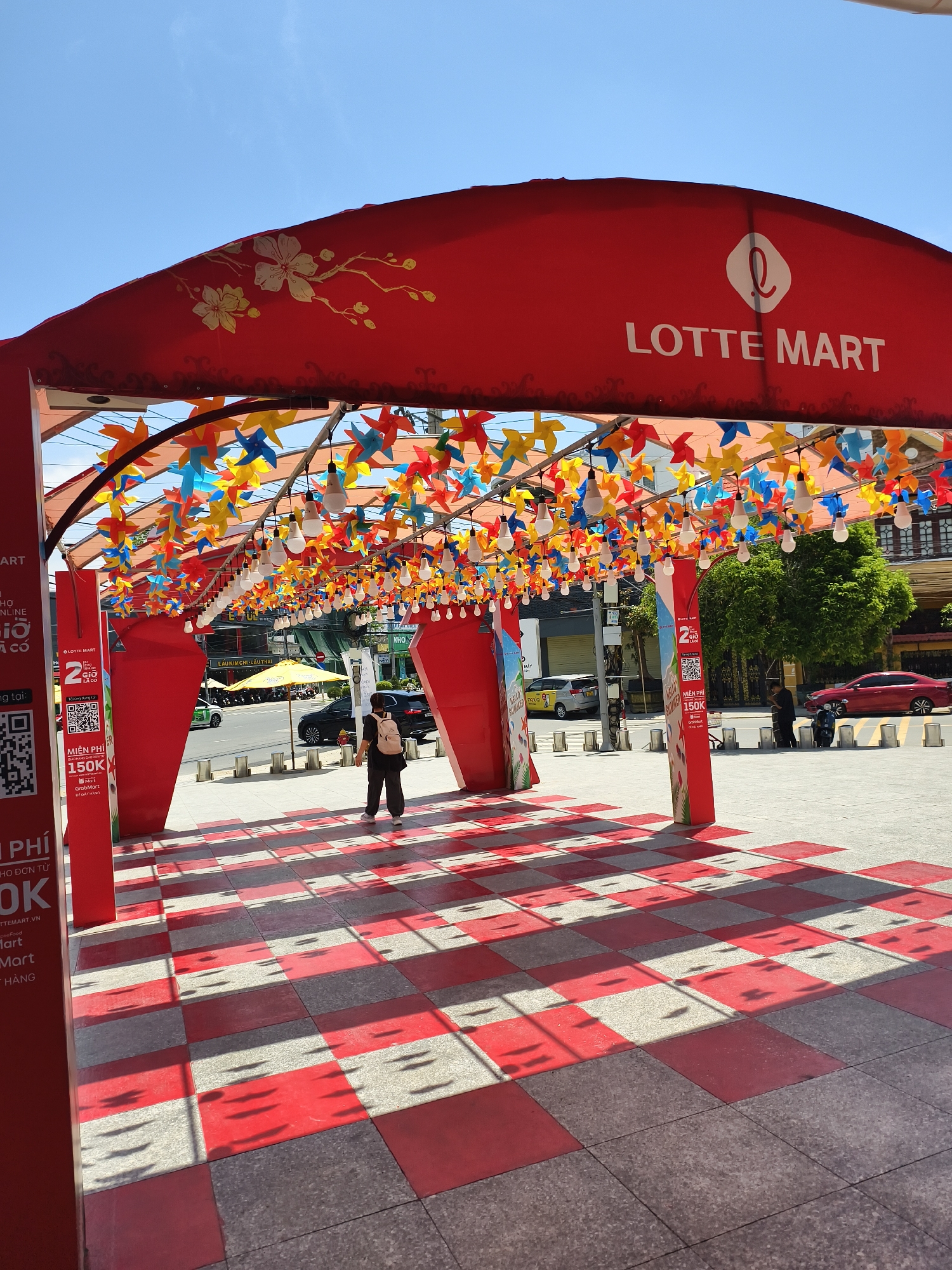 This image captures the decorated outdoor entrance of a Lotte Mart building in Tay Nha Trang Ward, Vietnam, on a bright, clear day. A prominent red archway with "LOTTE MART" in white lettering and a circular white logo is visible at the top center. The archway's underside is adorned with numerous strands of colorful paper pinwheels in shades of red, blue, yellow, and orange, interspersed with white, teardrop-shaped light bulbs, creating a festive ceiling. On the left side of the archway, a gold-colored floral design resembling cherry blossoms is painted on the red surface.
The ground beneath the archway features a checkerboard pattern of alternating red and grey square tiles. Sunlight casts distinct, elongated shadows from the overhead pinwheels and light bulbs onto these tiles, creating a patterned effect. Advertising banners and vertical red pillars with promotional QR codes and text in Vietnamese ("MIEN PHI 150K") flank the walkway.
One individual, dressed in dark attire with a light-colored backpack, is seen walking away from the viewer towards the street, positioned slightly right of the center under the archway. Beyond the archway, an active street scene is visible under a clear blue sky. This includes a two-lane road with several parked and moving vehicles, specifically a red sedan, a silver car, and multiple motorcycles. A yellow umbrella is open near the roadside, and green trees and multi-story buildings line the background.