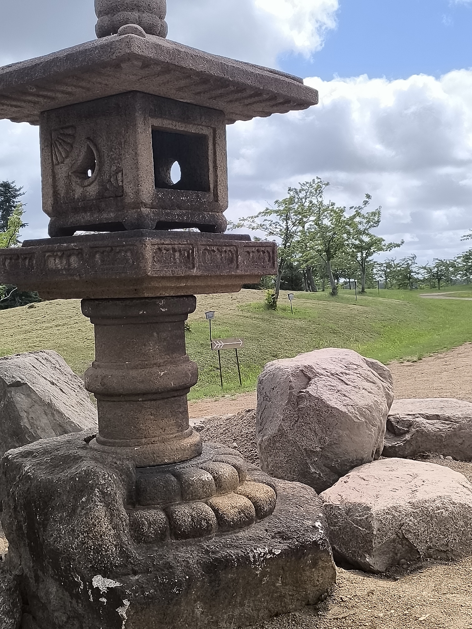 The image depicts a weathered stone lantern in what appears to be a park or garden setting. The lantern is a multi-tiered structure, with the main body being a square box with openings on each side, featuring decorative carvings. It rests on a decorative pedestal, which in turn sits on a rough-hewn stone base. Several large, rough-textured boulders are scattered around the base of the lantern. The background consists of a gently sloping grassy hill dotted with trees, and a pathway winding through it. The sky is partly cloudy with patches of blue. The overall scene is naturalistic and serene. The location is identified as Mažučiai, Lithuania.