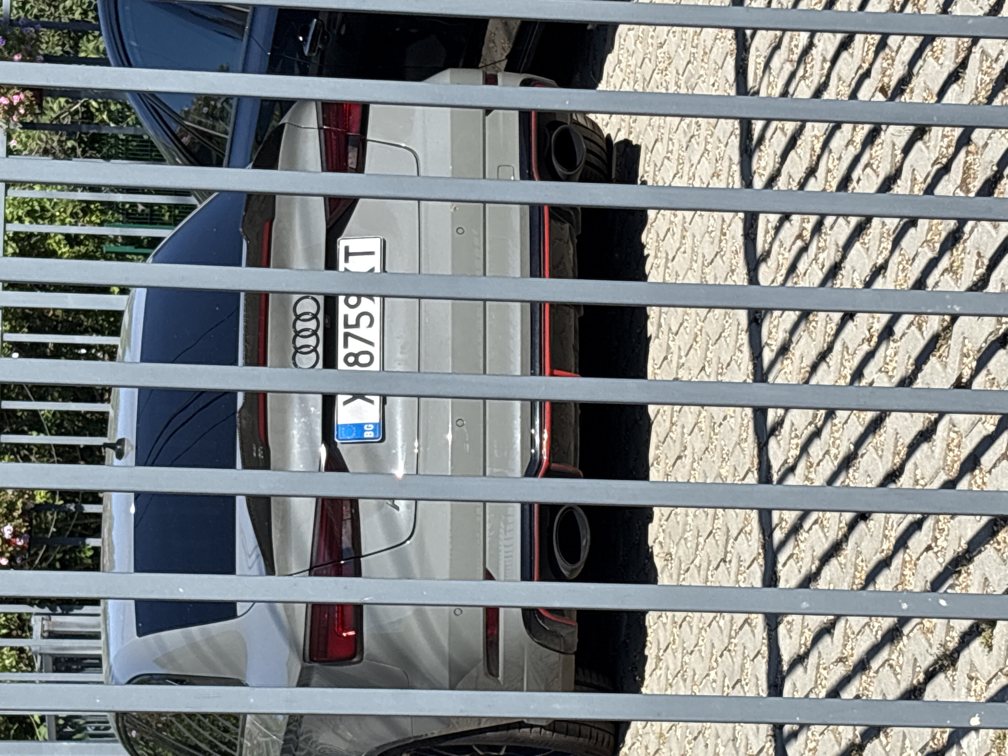 The image shows the rear of a light gray Audi sedan, partially obscured by a metal fence. The license plate is visible, displaying "BG X 8759 T". The car has distinctive red accents on its rear diffuser and taillights. Behind the Audi, a dark-colored car is parked. The ground is paved with interlocking concrete blocks, and shadows from the fence and foliage are cast across it. The scene appears to be in an outdoor parking area, possibly a private lot, given the fence. The location is specified as Varna, Bulgaria.