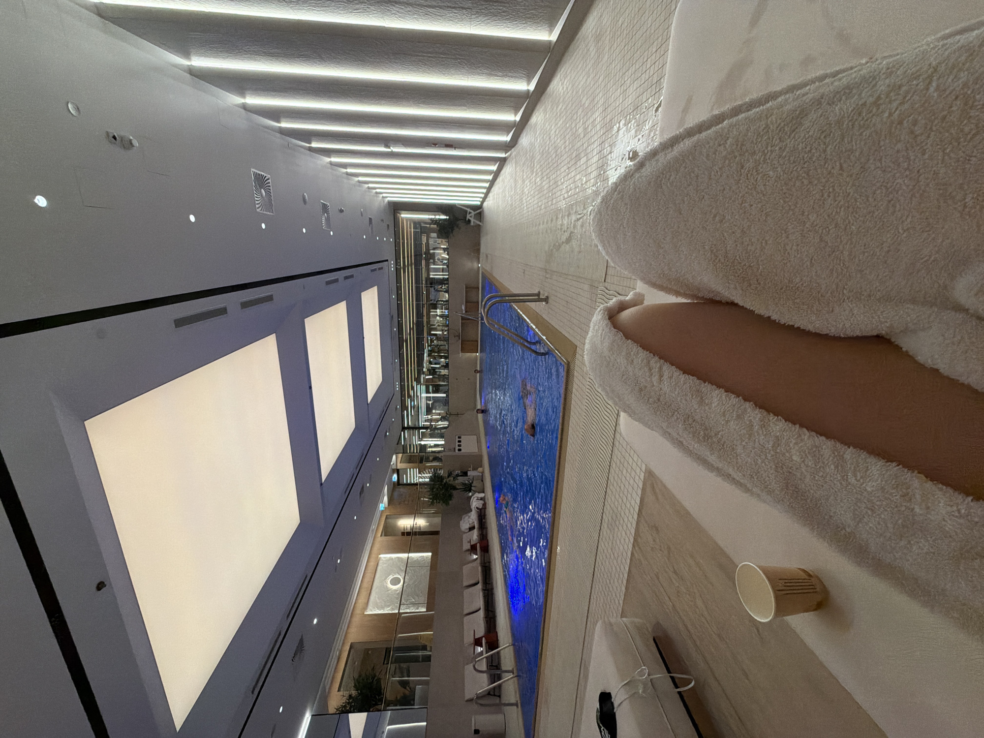 An indoor swimming pool with swimmers. A person in a white robe is sitting near the pool. A paper cup is on the ledge of the pool. The ceiling is white and lit. The location appears to be Unknown, Unknown.