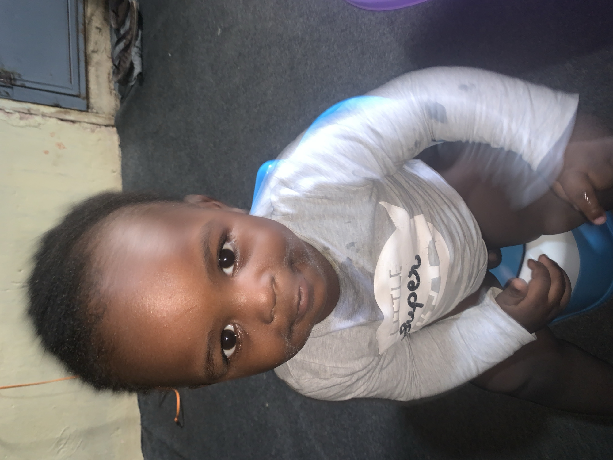 A dark-skinned toddler is seated on a blue potty training toilet. The child is wearing a light gray long-sleeved shirt with a graphic on the front that reads "Little Super". The background appears to be a room with a light green wall, a gray carpeted floor, and a dark blue door with a metal handle. There are also some shoes and other objects visible in the background, near the door. The overall lighting suggests the image was taken indoors, possibly in a dimly lit environment.