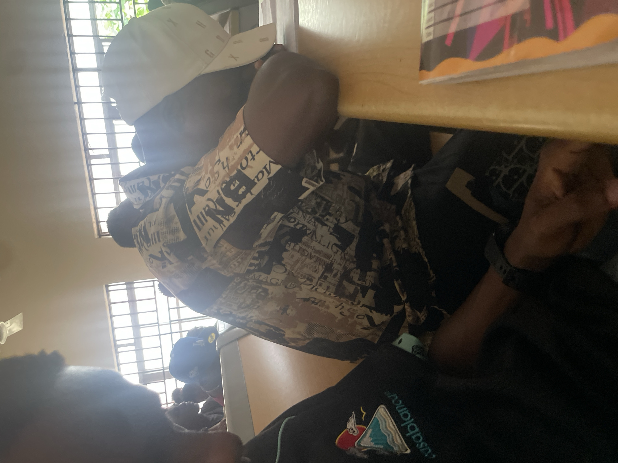 The image shows a group of young men in a classroom setting in Ihiagwa, Nigeria. The lighting suggests it might be daytime, with sunlight streaming in through barred windows. In the foreground, a person wearing a black shirt with a "Casablanca" logo is partially visible. To their right, another person is asleep or resting their head on their arms, wearing a patterned shirt and a white cap. The classroom appears to have standard wooden desks and chairs. The overall atmosphere seems relaxed, possibly during a lecture or study session where some individuals are taking a break.