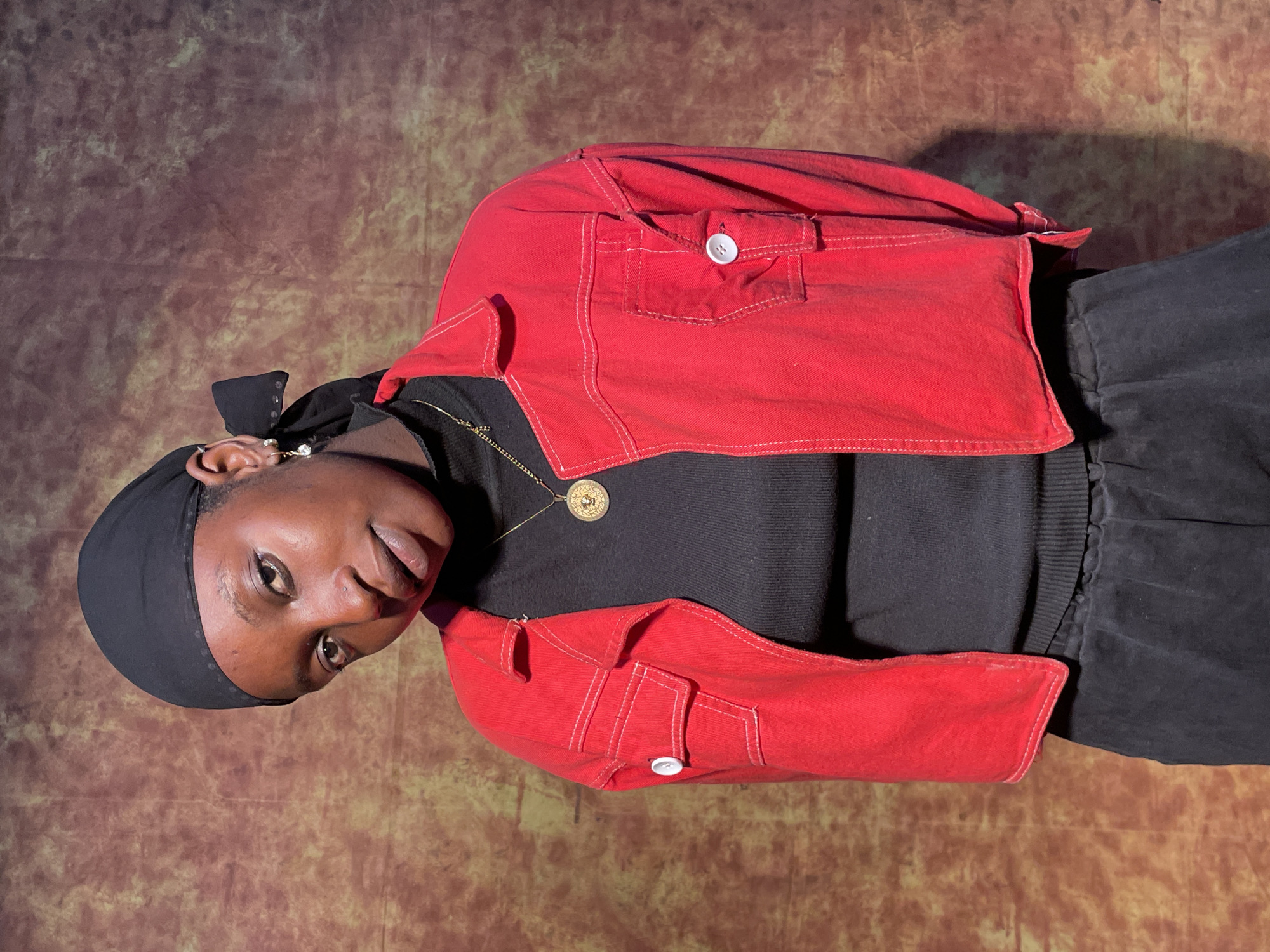 A person with dark skin and short hair covered by a black headscarf is wearing a black sweater and a short red jacket with white buttons and stitching. They are also wearing a gold necklace with a circular pendant. The background is a mottled brown and tan color. The location is unknown, as no details related to Barakin Akawo, Nigeria are visible in the image.