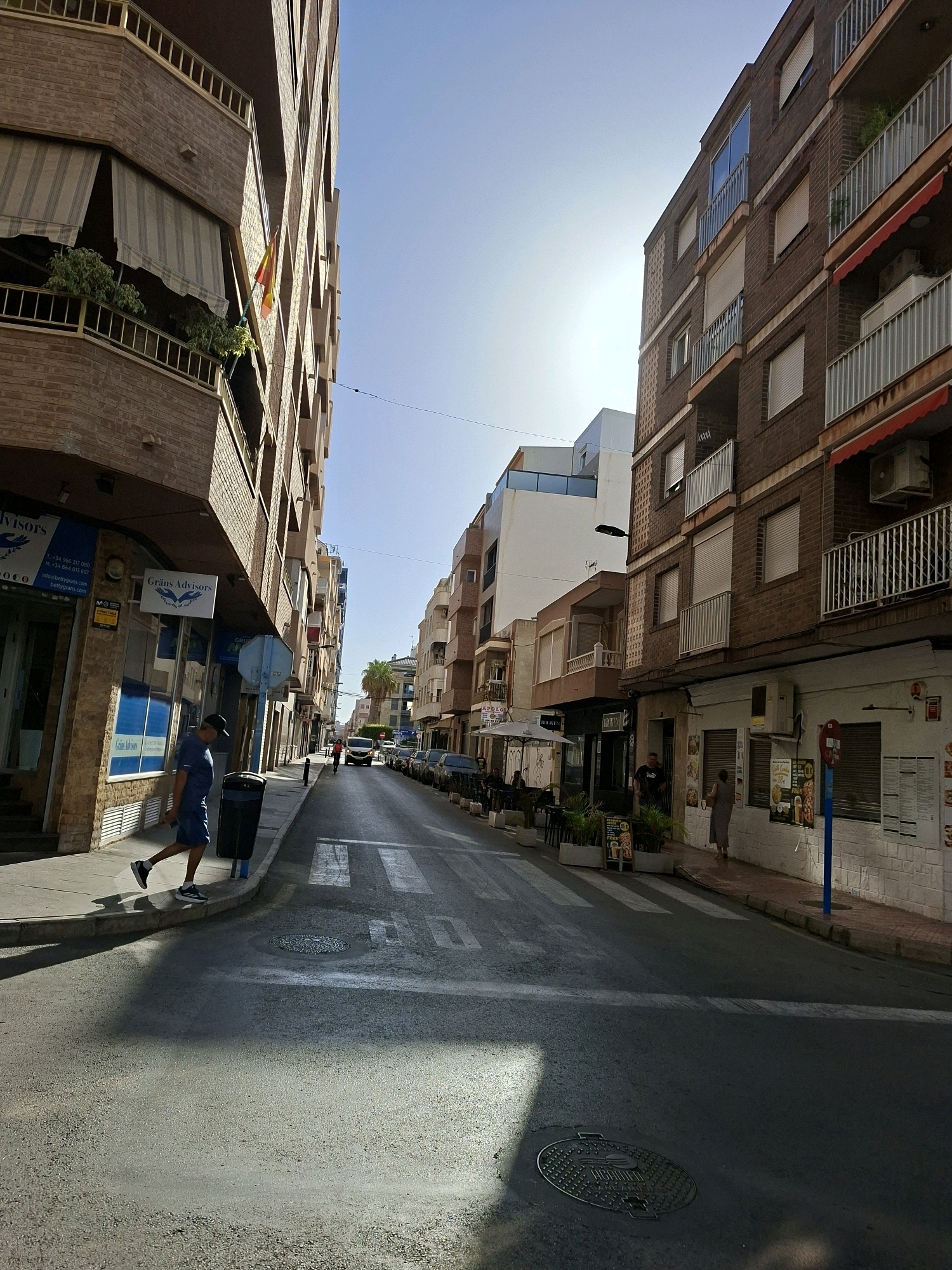A street view in Torrevieja, Spain, shows buildings lining both sides of an asphalt road with white crosswalk markings. On the left, a multi-story brick building has balconies with awnings and plants, and a Spanish flag is visible on one balcony. A man in a blue t-shirt and shorts walks on the sidewalk next to a black trash bin. Businesses with signage like "Grans Advisors" are located on the ground floor. The right side of the street also features multi-story buildings with balconies and awnings. Several cars are parked along the right side of the road, and one orange vehicle is driving down the street. A palm tree is visible in the distance. The sky is clear and bright, suggesting daytime.