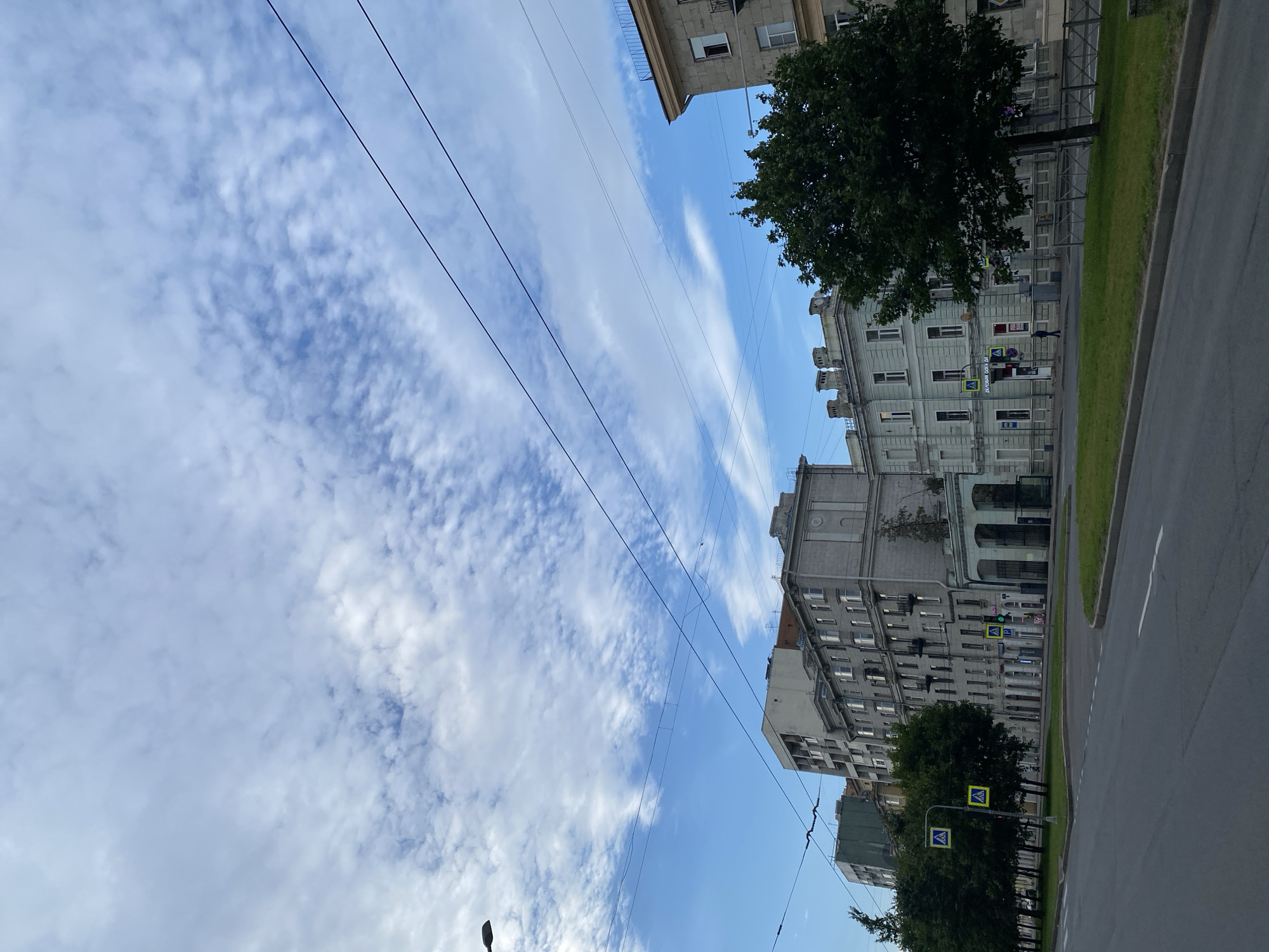 The image shows a street scene in Saint Petersburg, Russia, with several buildings lining the left side of a wide, grey asphalt road. The sky is a bright blue with scattered white, wispy clouds. Overhead, power lines run horizontally across the frame.
On the left side of the road, there is a row of trees casting shadows. Beyond the trees, several multi-story buildings are visible. The buildings are primarily made of grey stone and concrete, with varying architectural styles. One building in the foreground on the left is lighter in color, with many windows. Further down the street, there is a more ornate building with an arched entryway.
Several yellow pedestrian crossing signs with black figures are placed along the sidewalk. A few people are visible walking on the sidewalk in the distance. The overall impression is of a typical urban street on a cloudy but bright day.