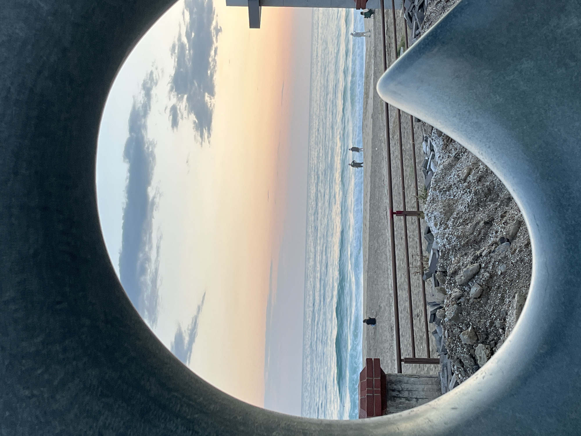The image depicts a beach scene in Alanya, Turkey, viewed through a circular architectural structure. The sky is visible with clouds and a gradient of sunset colors. The sea is adjacent to the beach, with waves breaking. On the beach, several people are visible at varying distances. A barrier separates the beach from a rocky, raised area. Parts of the structure framing the view are made of concrete.