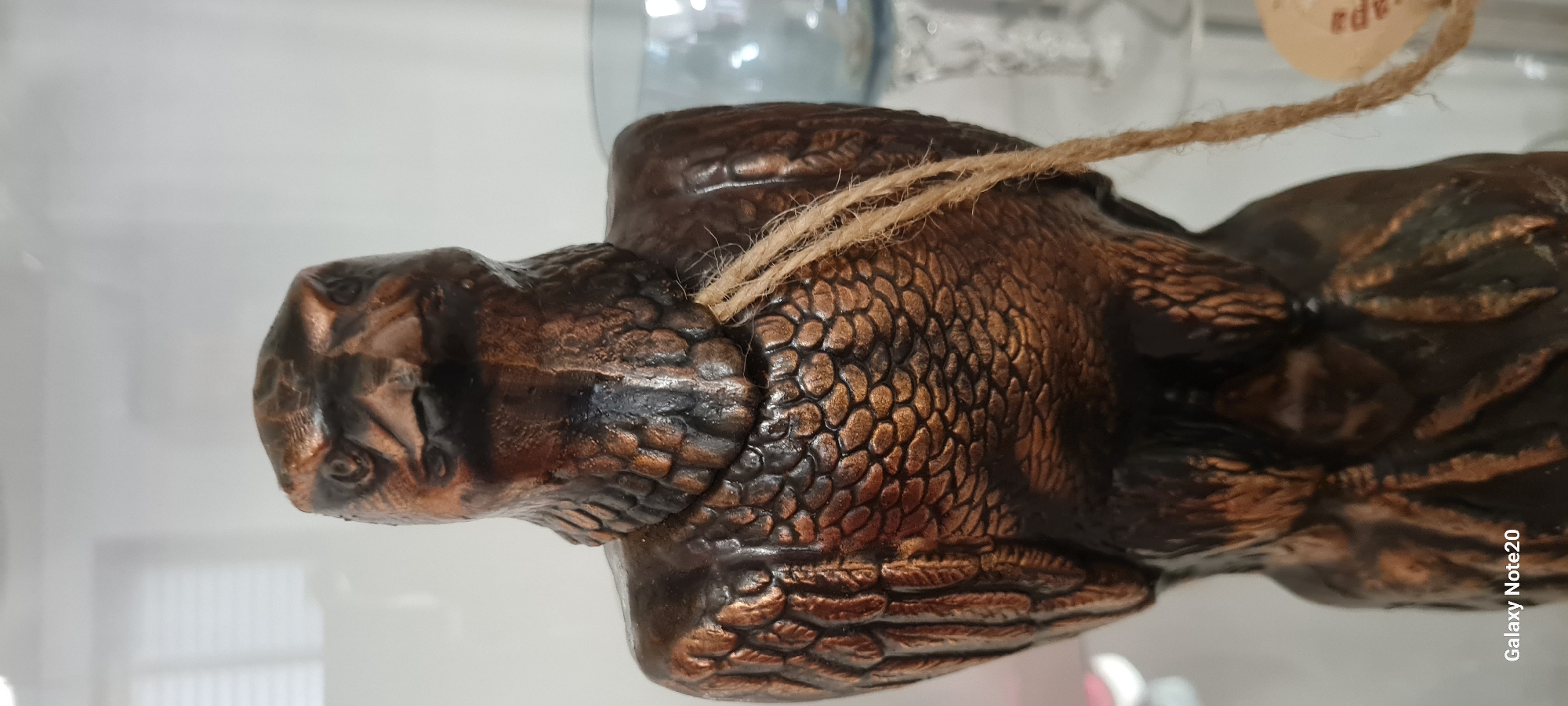 A close-up, eye-level shot shows a dark bronze-colored figurine of an eagle, viewed from the front. The eagle has detailed feathers on its body and wings, and its head is turned slightly to the left. A piece of natural twine is draped over the eagle's neck and hangs down its chest. The background is blurred and appears to be an indoor setting, possibly a shelf or display case, with hints of white and light blue objects. The bottom left corner of the image displays the text "Galaxy Note20." The image does not provide any information about the city of Holon, Israel.