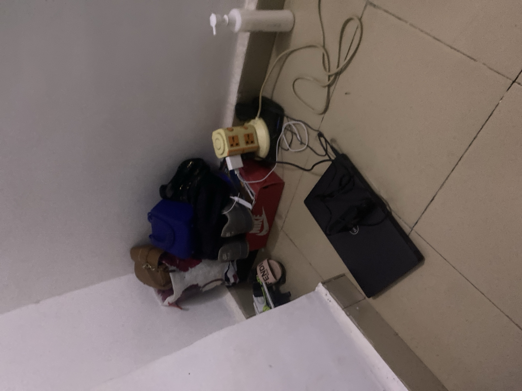 The image depicts a cluttered corner of a room, likely a bedroom, in Howayi, Nigeria. In the background, against a white wall, a pile of items includes a brown purse, a blue bag, and what appears to be a pair of brown shoes or boots. To the left of these, a white bag with a red and black pattern is visible. In front of the pile, a red shoebox with the Nike logo is present, containing a grey shoe. A pink slide with the "FENDI" logo is also placed on the floor.
In the foreground, a black laptop is open and positioned on the floor. Various electrical cords are scattered around the scene. A yellow power strip with multiple sockets is plugged into a black surge protector, and several white plugs are inserted into it. A white pump bottle, possibly containing soap or lotion, stands to the right of the power strip. The floor is tiled with light brown squares. No people are visible in the image.