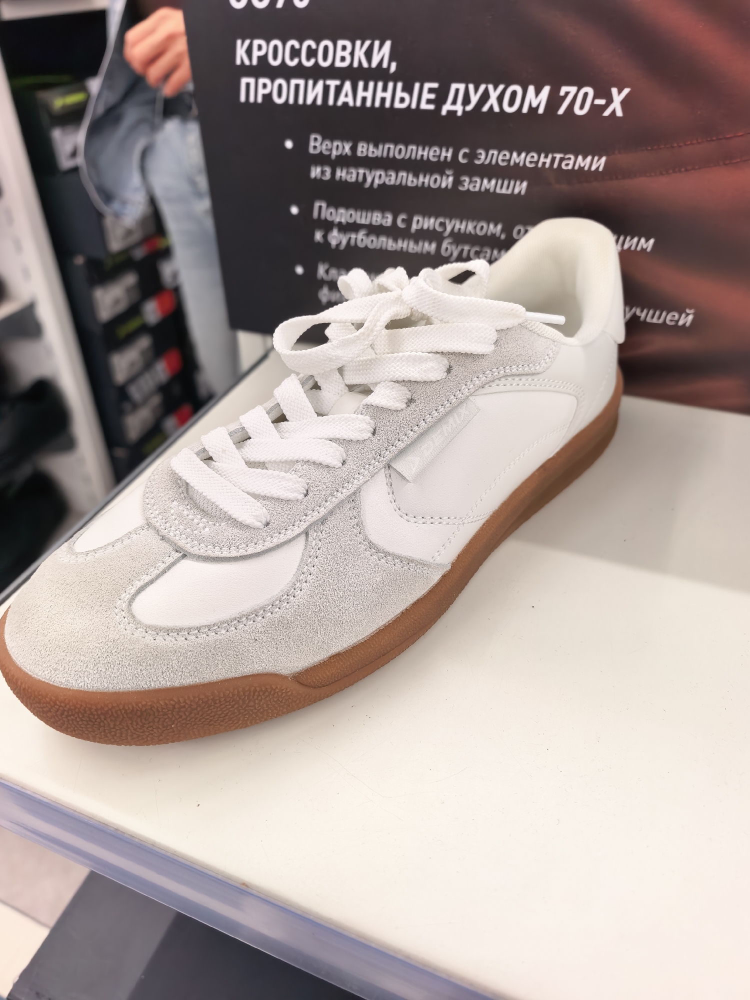 The image shows a close-up of a white and light gray athletic shoe with a gum sole. The shoe is resting on a white surface, possibly a shelf or display table in a retail environment. Behind the shoe, a dark sign with white text is visible, featuring bullet points describing product features in Russian. The sign mentions "КРОССОВКИ, ПРОПИТАННЫЕ ДУХОМ 70-Х" which translates to "Sneakers, infused with the spirit of the 70s." Other visible text on the sign refers to "Верх выполнен с элементами из натуральной замши" (Upper made with elements of natural suede) and "Подошва с рисунком, относящимся к футбольным бутсам" (Sole with a pattern related to football boots). The background on the right side of the image shows a gradient of red and brown tones. On the left side, blurred shelves with shoes are visible, suggesting a store setting. The overall impression is of a product display in a shoe store. Without additional information or context specific to Sterlitamak, Russia, it is impossible to determine the exact location within the city.