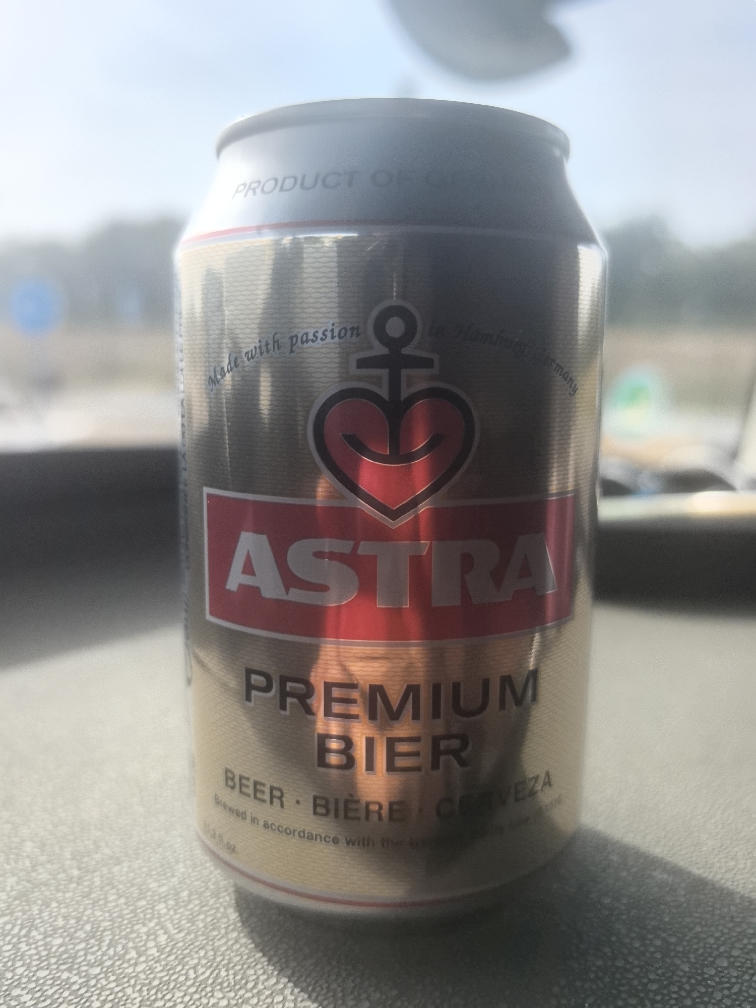 The image shows a close-up of a can of Astra Premium Beer, silver and gold in color, with a red band. The can is resting on a textured surface, possibly a table or countertop. The background is blurred, showing a bright, possibly outdoor scene with foliage and sky. There is a shadow cast on the surface from the can. The text on the can is legible, indicating "Astra Premium Bier" and other brewing information. No people are visible in the image. The location in Ciry-le-Noble, France is not determinable from the visual information provided.