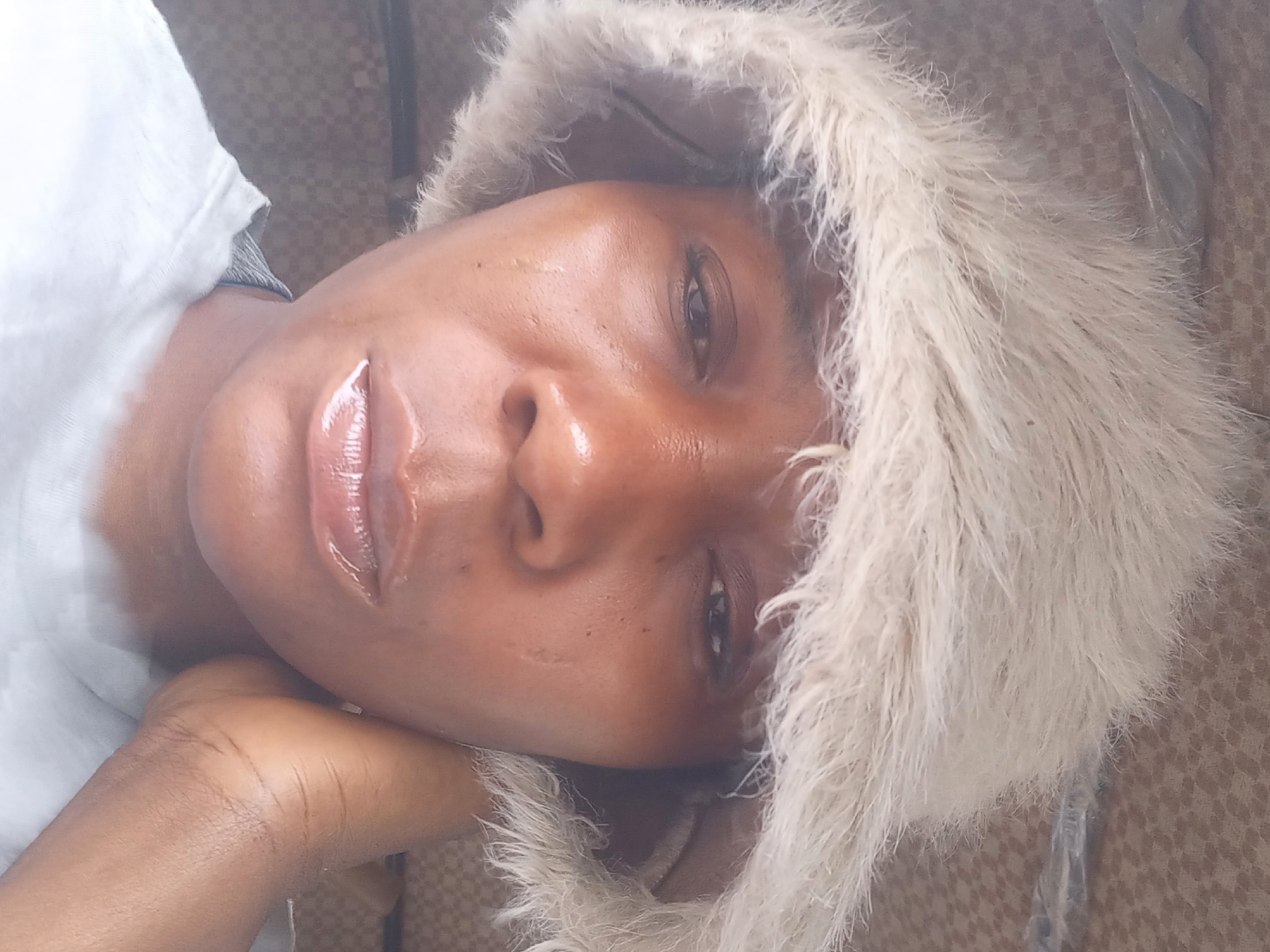 The image is a close-up selfie of a Black woman wearing a fluffy, light-colored hat. Her skin appears slightly oily, and she has on lip gloss. She is resting her right hand on her jawline, and her left hand is not visible. The background is out of focus but appears to be a textured surface, possibly fabric. The lighting suggests an indoor environment. There are no discernible elements that would allow for the determination of the city of Jos, Nigeria, as the location.