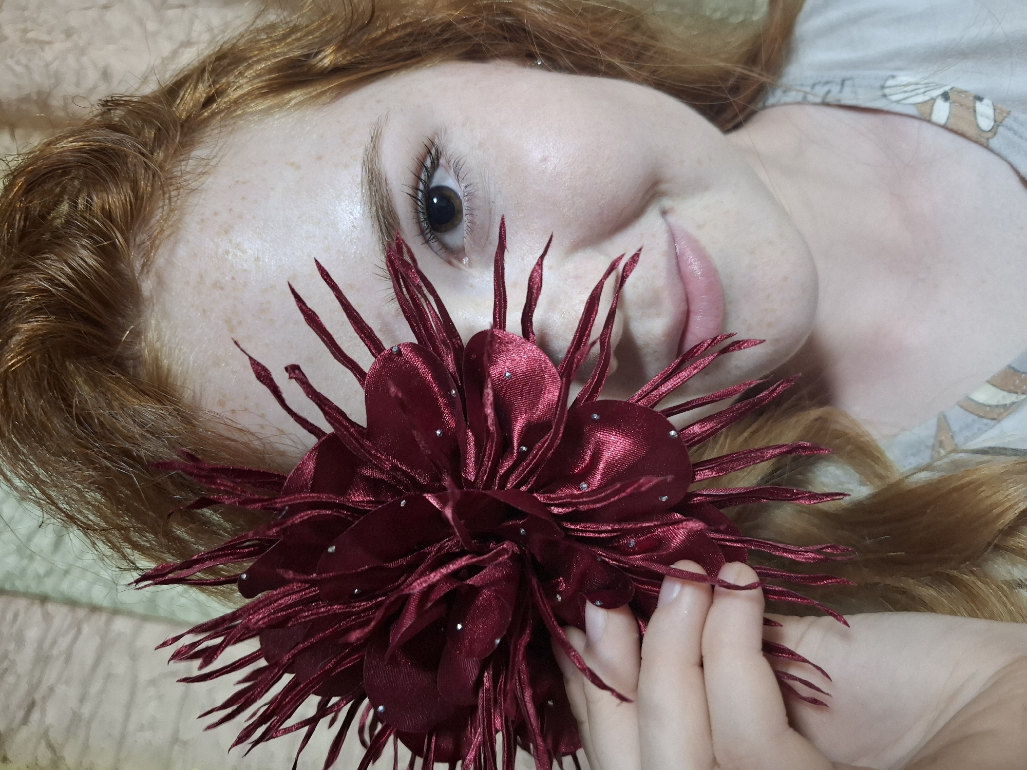 A fair-skinned woman with red hair is lying down, holding a large, red flower in front of her face. The flower is ornate with many layers of petals and small, shiny embellishments. The woman's left eye is visible, and she has freckles on her face. Her lips are slightly parted. She is wearing a light-colored shirt with a pattern on the sleeve. The background is a light, neutral color. The location is not discernible from the image.