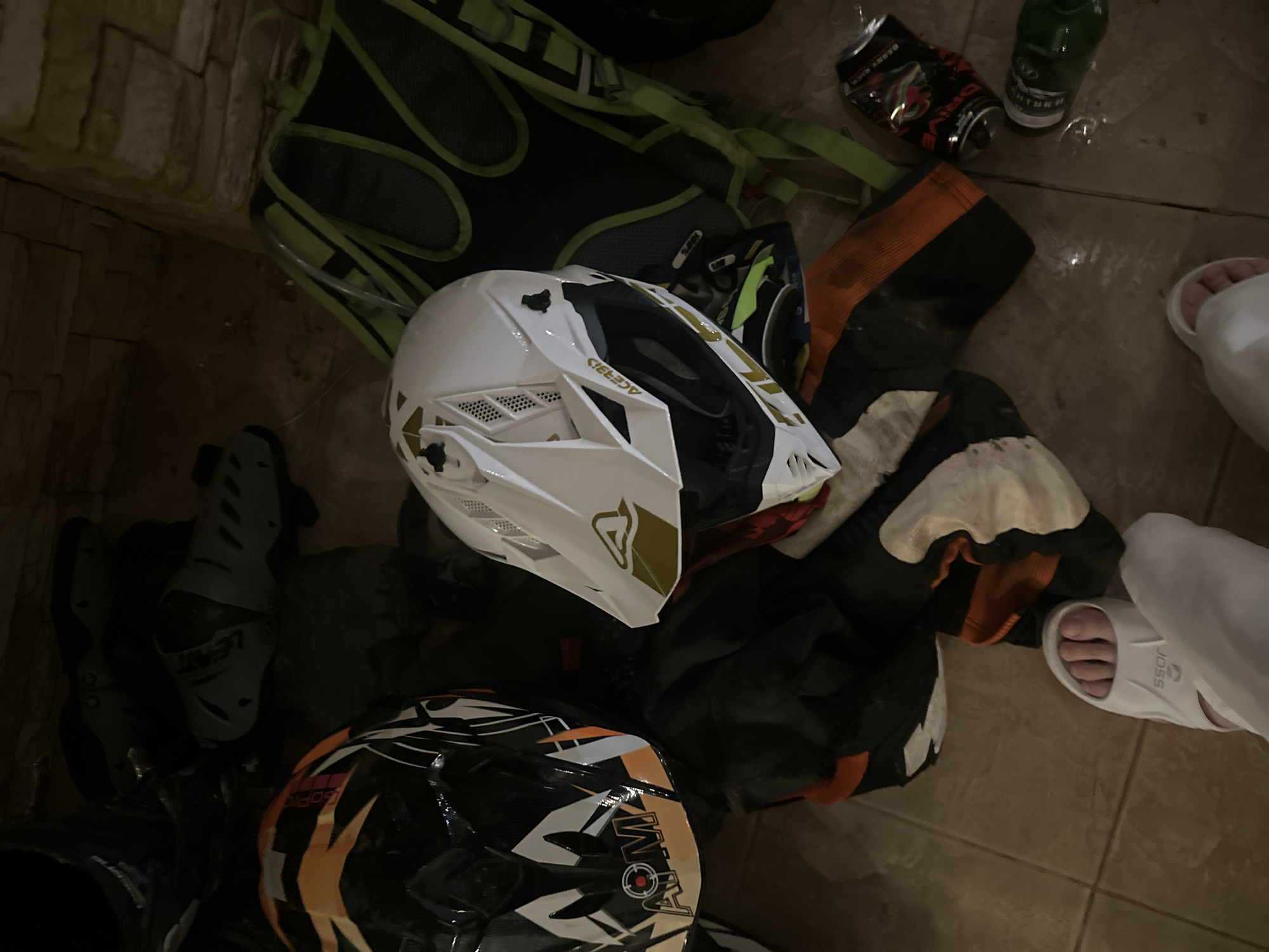 The image depicts a collection of motocross gear and associated items scattered on a tiled floor, likely in a residential or storage area in Orekhovka, Russia. Two motocross helmets are visible: one black and orange with "AOM" branding, and another white with gold accents and "Acerbis" branding. Alongside the helmets are protective knee pads, one with "Leatt" branding. A bright green and black backpack is present, as are sections of motocross pants with orange and black detailing. Additionally, a white slide sandal worn by a person whose feet are visible in the lower part of the frame is present, along with a crushed energy drink can and a partially visible green bottle. The environment suggests a casual or post-activity scene.
