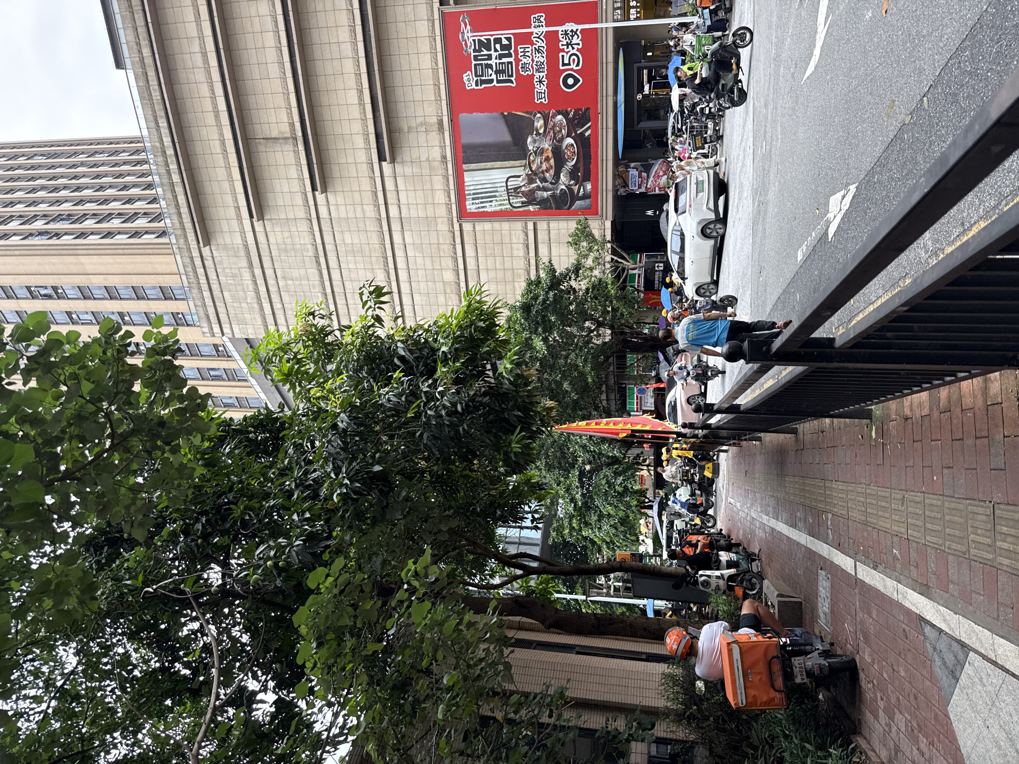 In Haizhu District, China, a street scene unfolds with a sidewalk lined by trees and a brick building on the left. Several individuals on motorcycles are parked or moving along the sidewalk, some appearing to be delivery personnel with orange boxes on their vehicles. A large building with a beige facade and numerous windows dominates the background, with a tall, lighter-colored skyscraper behind it. A prominent red billboard displays an advertisement for a hot pot restaurant, featuring images of food and Chinese text. On the street, cars and more motorcycles navigate the asphalt, with some pedestrians carrying umbrellas, suggesting overcast weather. A black metal fence separates the sidewalk from the street.