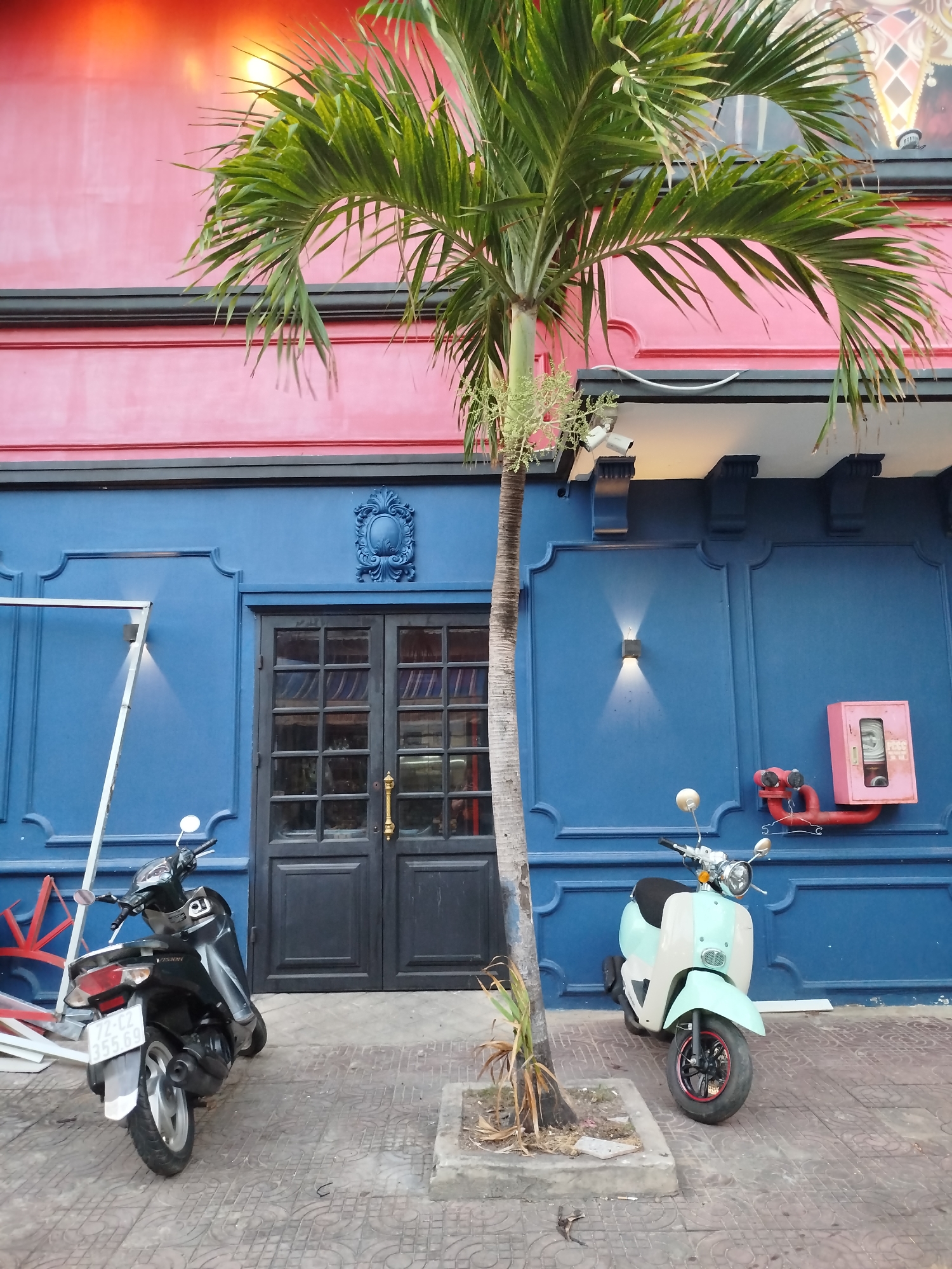 The image depicts a street scene in Ho Chi Minh City, Vietnam. The focal point is the exterior of a building with a two-tone facade: a red upper section and a blue lower section. In the center of the blue section are black double doors with glass panes, flanked by decorative molding and a carved ornament above the doors. A small palm tree grows in front of the building. Two scooters are parked on the sidewalk in front of the building. The one on the left is dark-colored, and the one on the right is a light blue and white. A red fire hydrant and a pink fire hose box are mounted on the blue wall to the right of the doors. Additional architectural details include sconces and decorative black corbels under a white overhang. The sidewalk is paved with gray tiles. The scooters partially obscure the view of the storefront and the sidewalk in front of the building. A white metal frame with some red pieces is to the left of the left scooter.