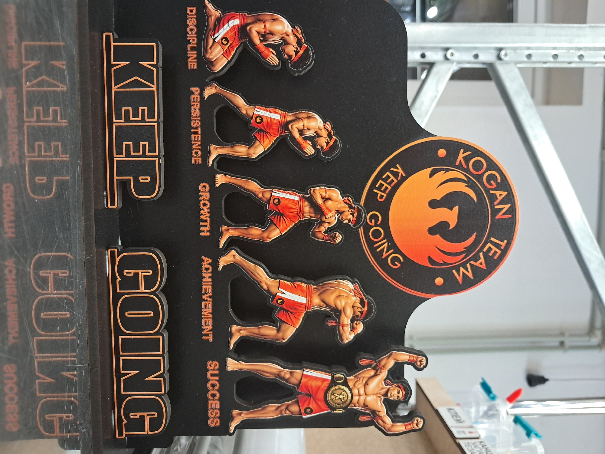 The image displays a black, three-dimensional motivational display related to martial arts. The display features a circular logo at the top with an orange phoenix and the text "KOGAN TEAM KEEP GOING" in orange lettering. Below the logo, there are five figures of a Muay Thai fighter in various stages of training, progressing from left to right. The figures are depicted in orange shorts and headbands. Beneath each figure is an orange word representing a stage of progress: "DISCIPLINE," "PERSISTENCE," "GROWTH," "ACHIEVEMENT," and "SUCCESS." At the bottom of the display, the words "KEEP" and "GOING" are presented as separate, large, three-dimensional orange and black letters, also with their reflections below. The background appears to be a generic indoor environment with metal shelving structures visible behind the display. The display is positioned on a reflective surface.