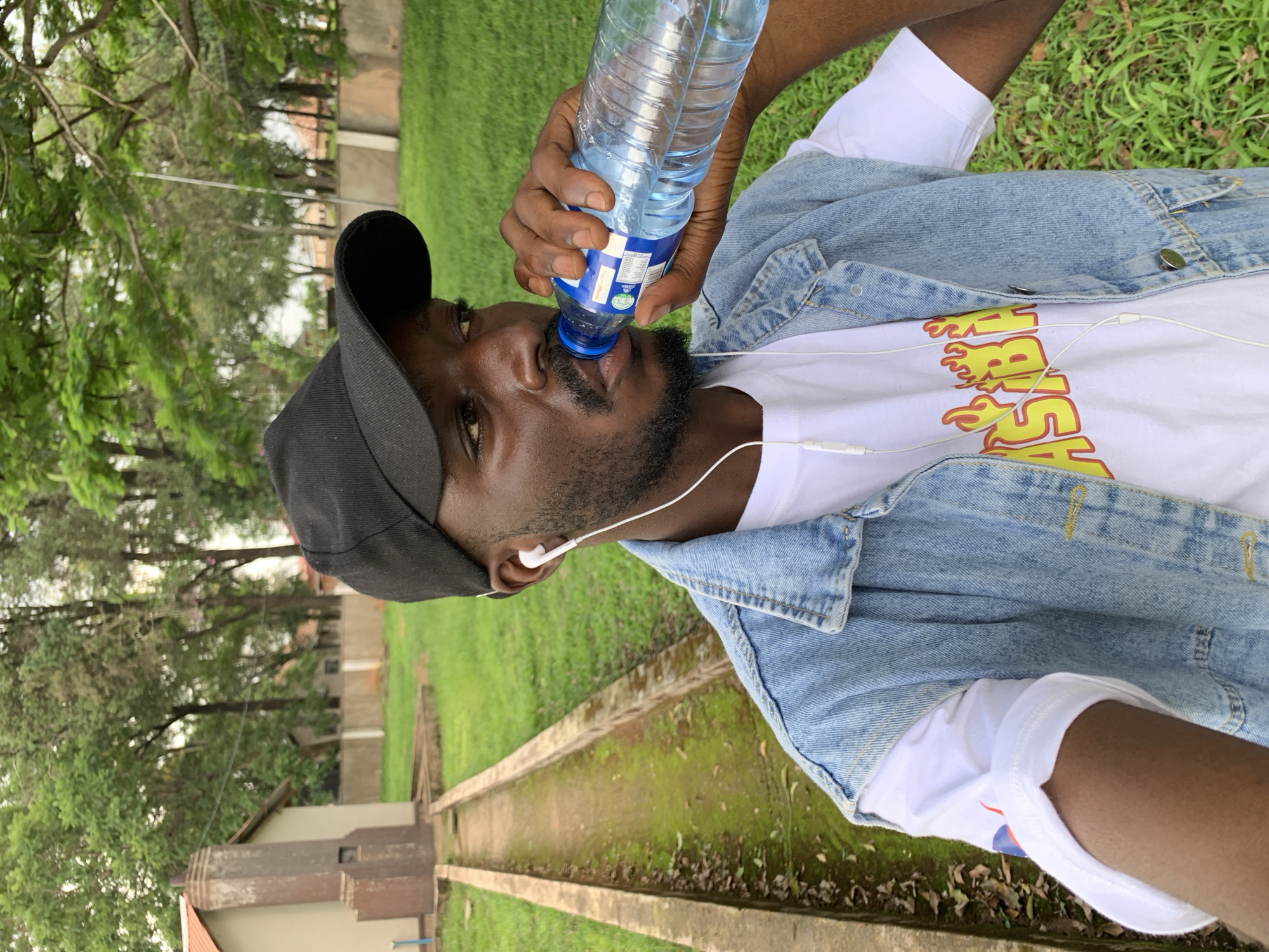 The media file shows a medium close-up shot of a Black man from the chest up, facing the camera. He is wearing a black baseball cap, a white t-shirt with a yellow and red flame-like graphic design, and a denim jacket. He is drinking from a clear plastic water bottle held in his right hand. White earbuds are visible, one in his ear and the wire trailing down his chest. The man has dark skin, a beard, and appears to be in his early to mid-twenties.
The setting is outdoors, with a background of green grass, trees with lush foliage, and a concrete pathway or drainage system. In the distance, a building or structure is partially visible, suggesting an urban or semi-urban environment. The lighting suggests it is daytime, possibly overcast or shaded by the trees. The overall mood is casual and relaxed. The graphic on the t-shirt is difficult to fully decipher due to the angle and resolution, but the words "FIRE" or "SMOKE" are suggested by the lettering. No other visible text is clear. The scene could be a park, university campus, or residential area in Jos, Nigeria.