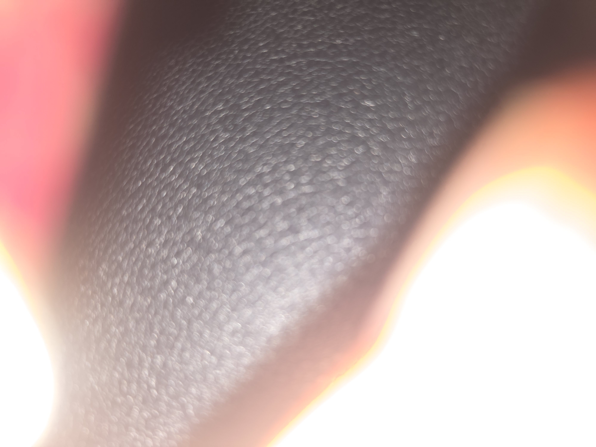 The image shows a close-up, out-of-focus view of a textured black surface, possibly leather or similar material. A blurry pink or red area is visible on the left side of the frame. The right and bottom sections are overexposed, appearing as bright white with orange and yellow light flares. The location in the city of Pintomakmur, Indonesia is irrelevant because the image does not depict any geographical landmarks or specific features indicative of that location.