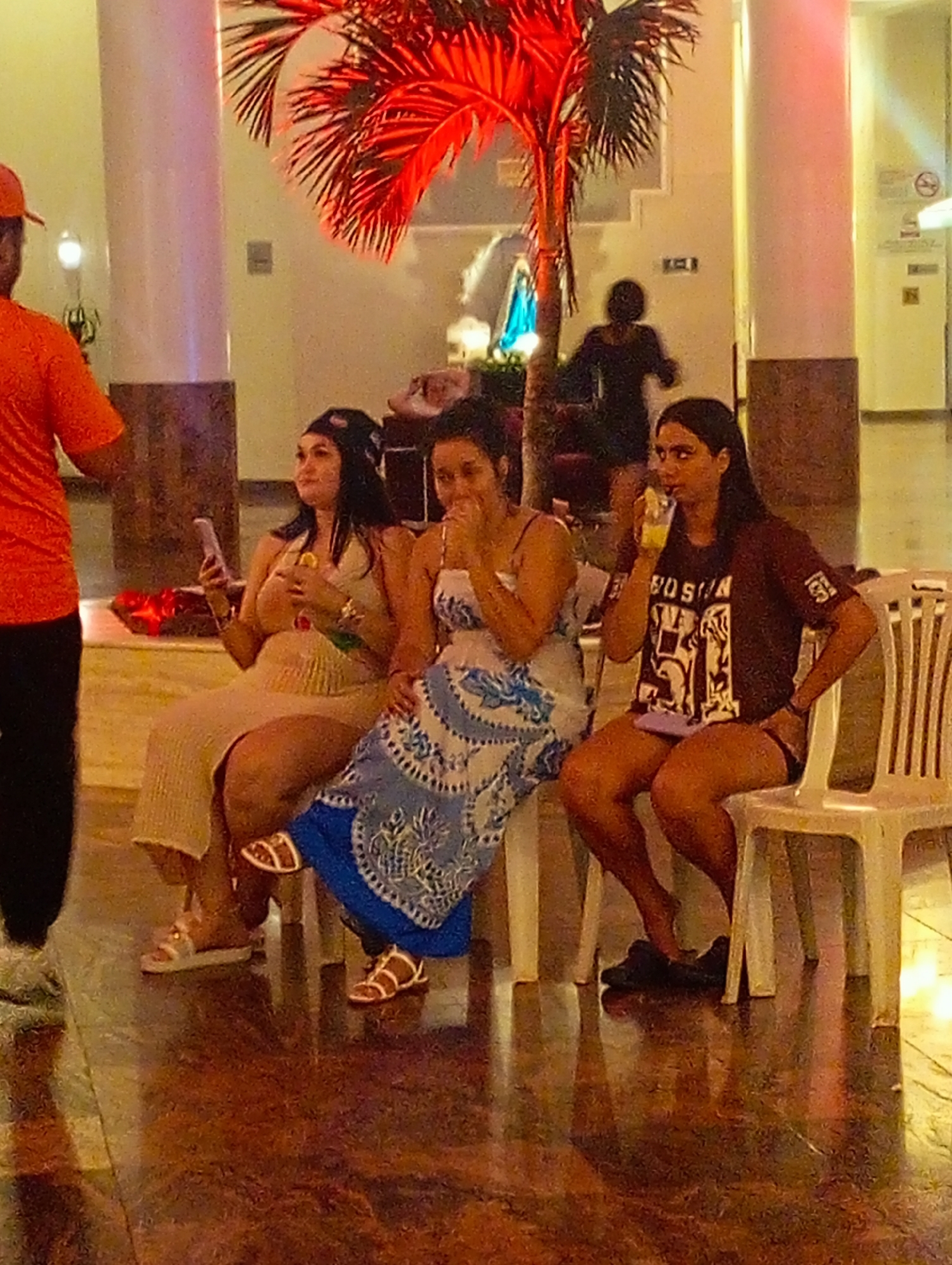 In Pedro González, Venezuela, a gathering takes place within an indoor location featuring tile flooring, likely a hotel or resort lobby given the presence of pillars and a decorative red palm tree. Three women are seated on white plastic chairs; one is drinking from a glass, another appears to be reacting to something, and the third is holding a smartphone. An individual in an orange shirt partially obscures the left side of the frame. A person in dark clothing is further back behind the artificial tree. There is a no smoking sign in the background.