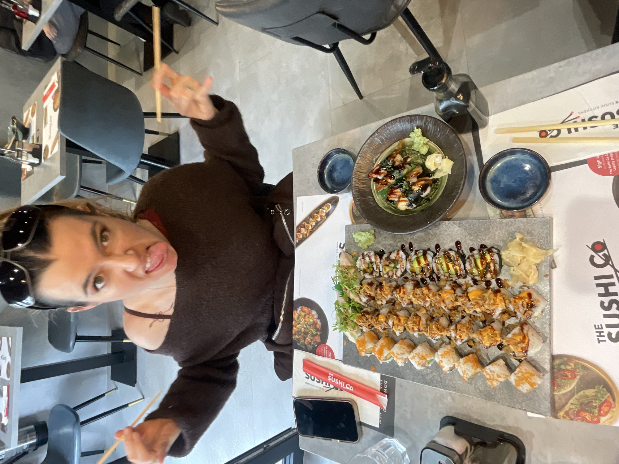 Here is a detailed description of the image:
A person is seen posing with chopsticks in front of a table laden with food. The food includes a plate of sushi with various toppings, a bowl of what appears to be some kind of salad or greens, and a small dish with sauce. There is also a bottle, presumably containing soy sauce or another condiment. On the table are items related to "The Sushi Co", indicating that this is a restaurant or takeout from that establishment. There's also a mobile phone.
The person is wearing a dark-colored sweater and sunglasses. They are sticking out their tongue, suggesting a playful or silly pose.
The image suggests that it was taken at "The Sushi Co" a sushi restaurant in the city of London, United Kingdom, given the reference to "THE SUSHI CO" and "SUSHI.CO", and the phrases "TE SUSHI KITCHENS" on the printed materials.