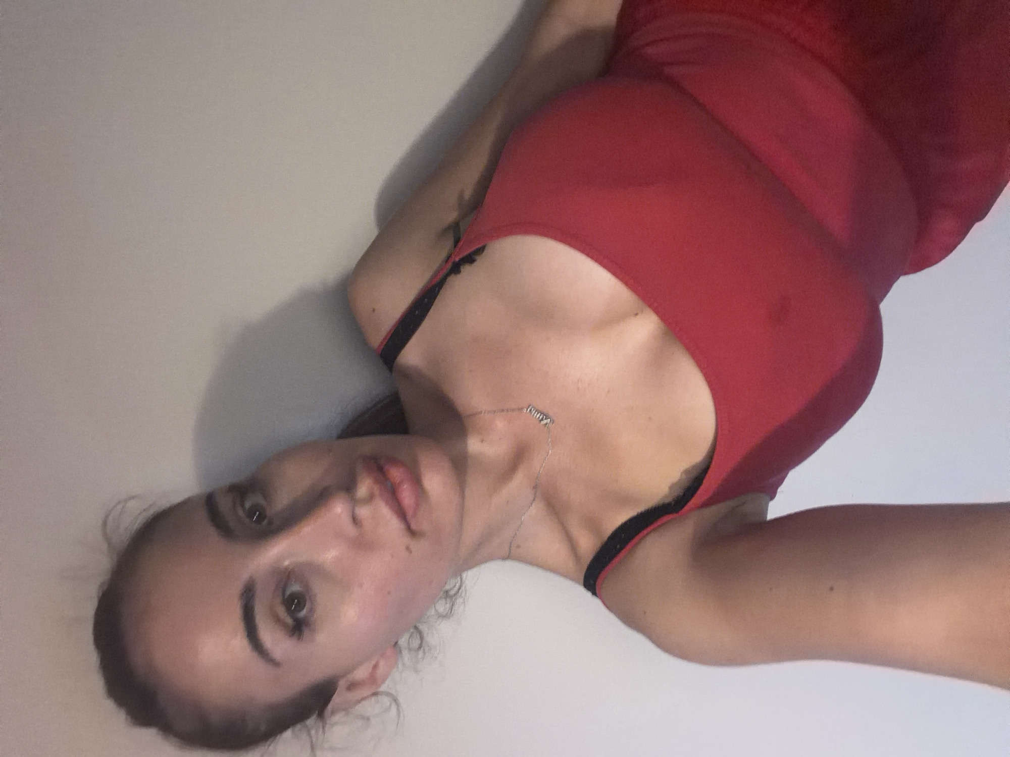 A woman is lying down against a wall, her body oriented vertically in the frame. She is wearing a red top with black straps, and a thin silver necklace with a charm is visible around her neck. The lighting suggests an indoor setting, possibly at home, with a soft shadow cast on the wall behind her. Her facial expression is relaxed, and her gaze is directed towards the camera. The overall scene is simple and intimate, with no specific activity other than posing for the photograph. There is no visible text in the image. The location context provided is Tersefanou, Cyprus.