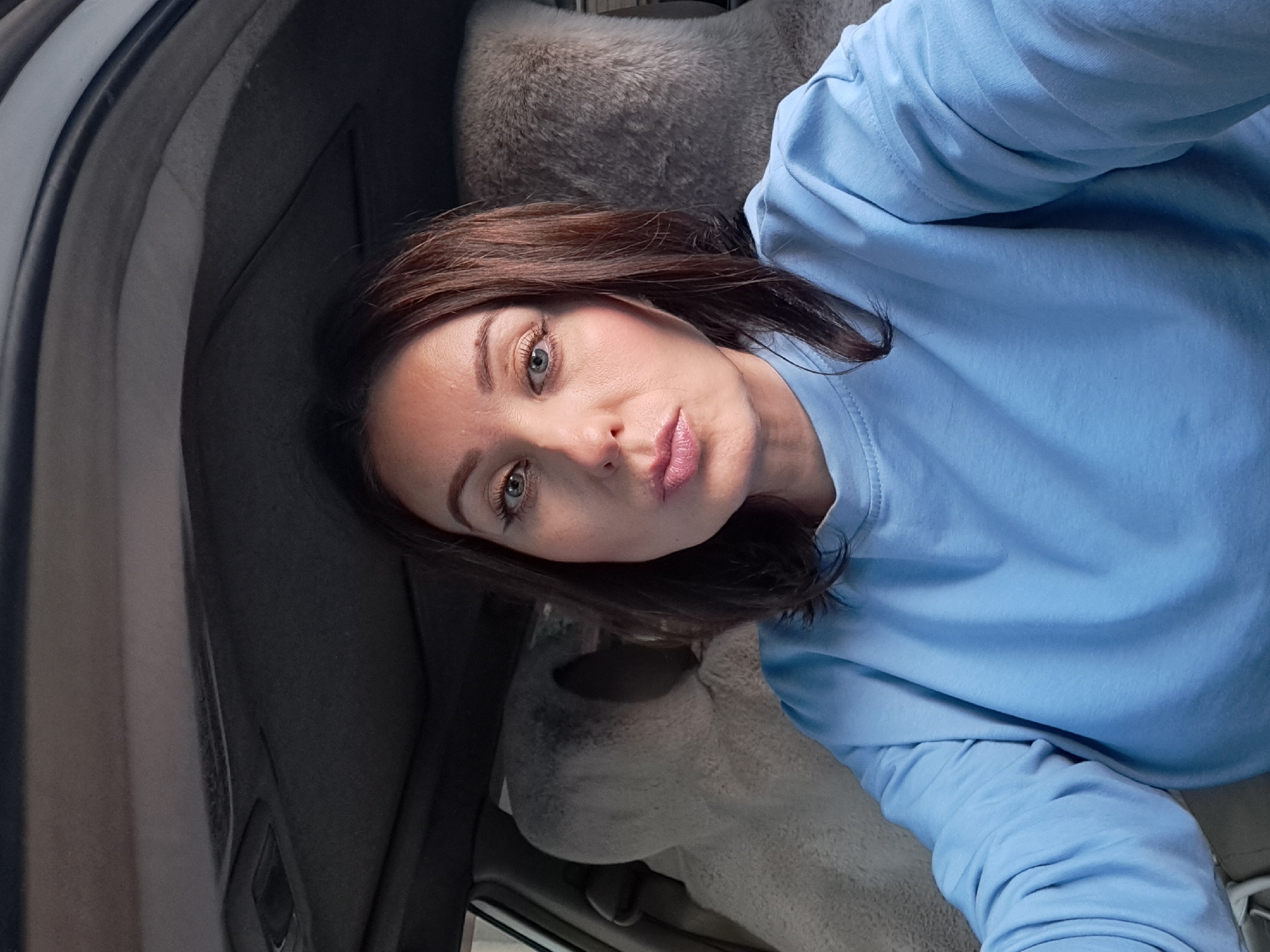 A woman with dark hair and light-colored eyes is sitting in the driver's seat of a car, looking at the camera. She is wearing a light blue sweatshirt. The interior of the car is visible, including fuzzy seat covers. The image appears to be taken from the front passenger seat. The background suggests an indoor location, possibly a garage or parking structure. No specific landmarks or indicators of the city of Irkutsk, Russia, are present in the image.