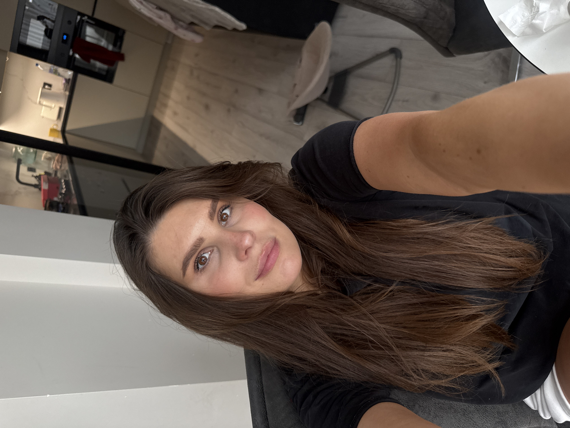 The image displays a selfie of a young adult female taken indoors, depicting a residential environment in Sofiivska Borshchahivka, Ukraine. The woman is seated, facing the camera with a slight smile, and her right arm is extended to hold the device. She has long, dark brown hair, brown eyes, and is wearing a black short-sleeved t-shirt. A portion of white clothing is visible below her black top.
The background shows a modern kitchen area to the right, featuring light-colored lower cabinets, dark countertops, and a built-in oven with a blue digital display. A red cloth hangs from the oven handle. On the countertop, a white kettle, a yellow mug, a water bottle, and other items are visible. The flooring is a light wood laminate. In the middle ground, a light-colored high chair or booster seat with metal legs is positioned. To the left of the woman, a plain white wall is visible. The scene is well-lit, indicative of an interior domestic setting.