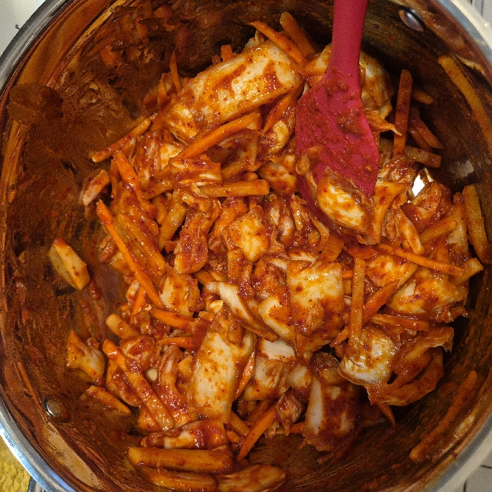 A stainless steel pot is filled with kimchi and shredded carrots, mixed with a vibrant red paste. A pink spatula is partially visible, suggesting the food is being mixed. The ingredients appear glossy and well-coated in the sauce. The pot's interior shows some wear and discoloration. The overall composition suggests preparation or mixing of the kimchi.