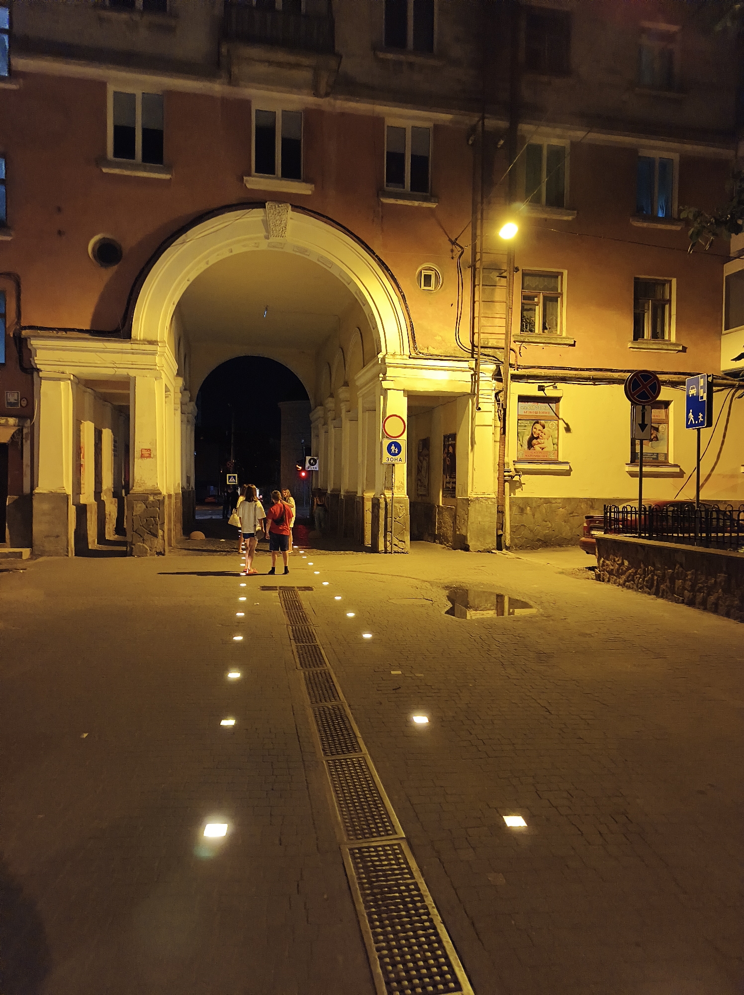 The image depicts a wide, illuminated pedestrian passage under an archway of a building in Ternopil, Ukraine, at night. The architecture features a large, arched opening supported by sturdy pillars. The facade of the building is a warm, terracotta color with multiple windows. The ground is paved with cobblestones and features a drainage grate running along the side. Small, square lights are embedded in the pavement, illuminating the path.
In the foreground, two individuals, a male and a female, are walking away from the camera through the passage. The male is wearing a red t-shirt and dark shorts. The female is wearing a white t-shirt and light-colored shorts, carrying a bag. They are the primary subjects in the immediate vicinity.
Further into the passage, a few other figures are visible, though less distinctly. The environment is urban, with streetlights casting a yellow glow and creating reflections on the wet pavement, indicating recent rain. Several signs are posted on the building walls, including traffic and informational signs. One poster is visible in a window on the right side of the passage. The overall scene suggests a quiet evening in a city center.