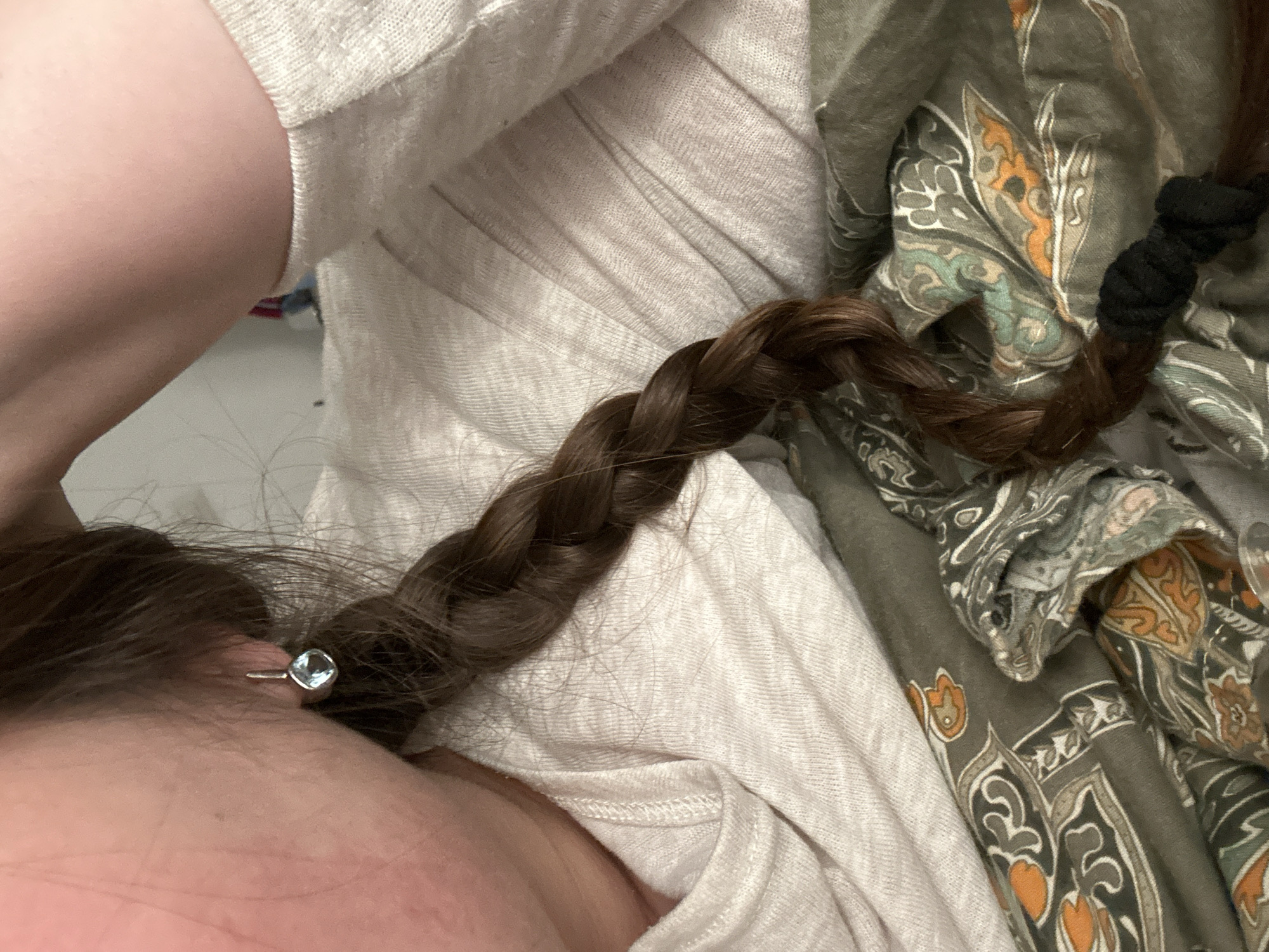 A close-up shot presents a partially visible person with light skin, a brown braided hair extension, and a small, square-shaped earring with a silver post. The braid is secured with a black hair tie. The person is wearing a light beige shirt with textured fabric. The background is a patterned cloth featuring shades of olive green, brown, and orange. This image does not have details that lead to Ramat Gan, Israel.