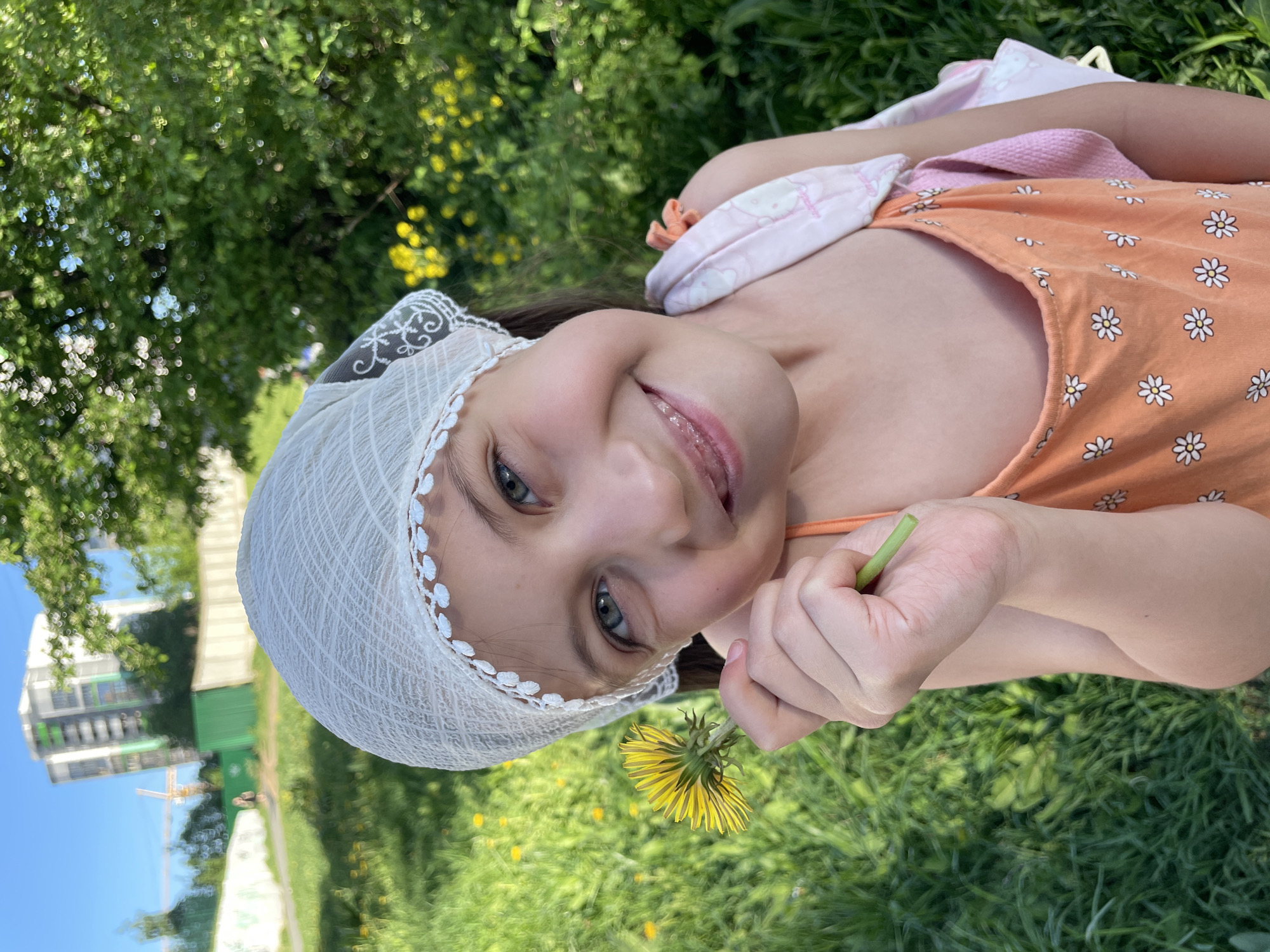 A young girl with a white headscarf is lying in a grassy field, holding a dandelion flower. She is wearing an orange dress with white flower patterns and a pink and white patterned shoulder bag. The background includes green grass and trees, with a building and construction crane visible in the distance. The photo appears to be taken on a sunny day. The location is in Izhevsk, Russia.