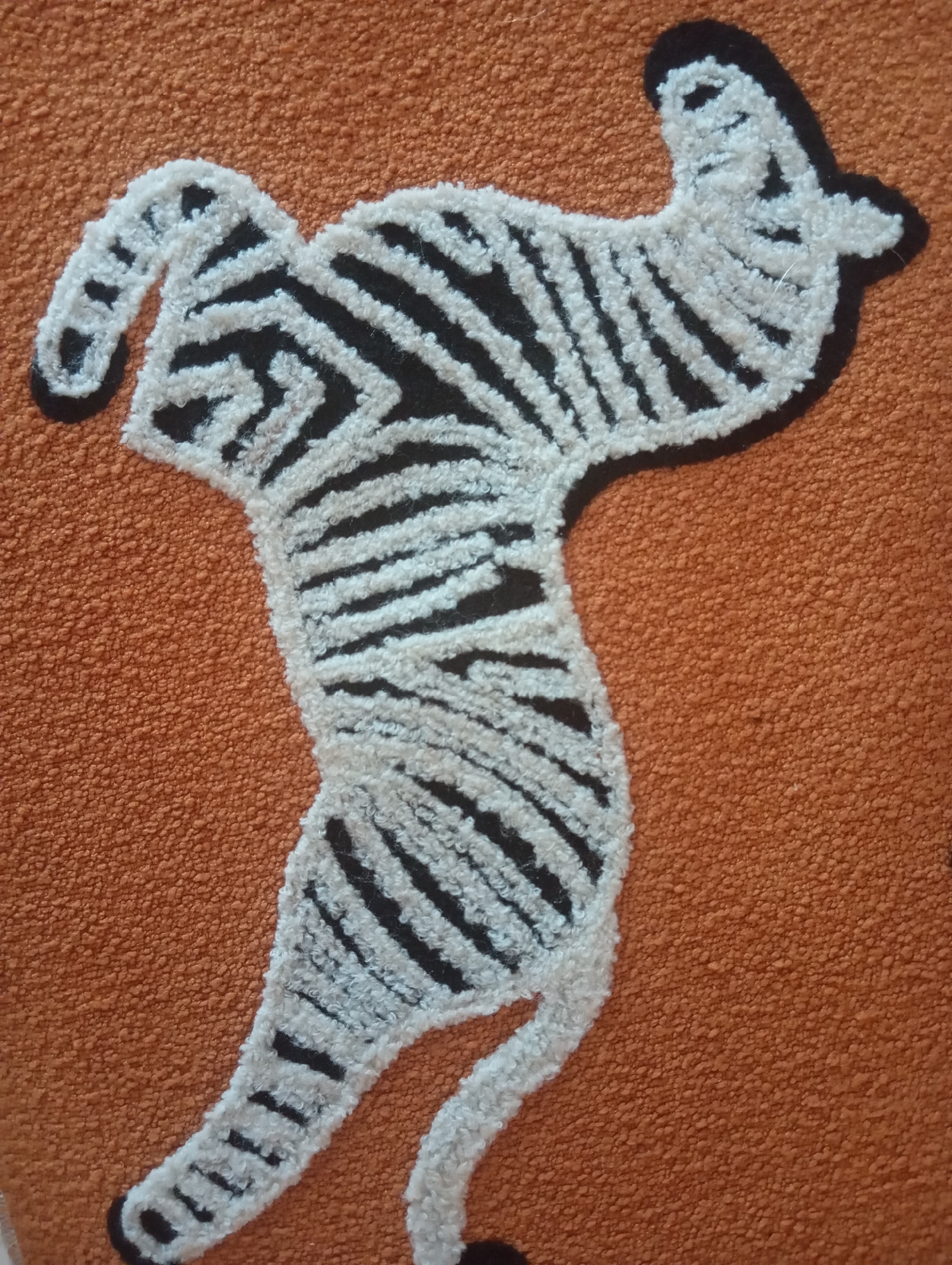 A close-up image shows a textured rug with a zebra design. The rug is a warm orange color and features a stylized zebra made of white and black yarn. The zebra's stripes are prominent, and its body is outlined in black. The texture of the rug is coarse, with many small, raised loops. The image is shot from a slightly elevated angle, focusing on the zebra design. There is no indication of the city of Karon, Thailand, or any people in the image.