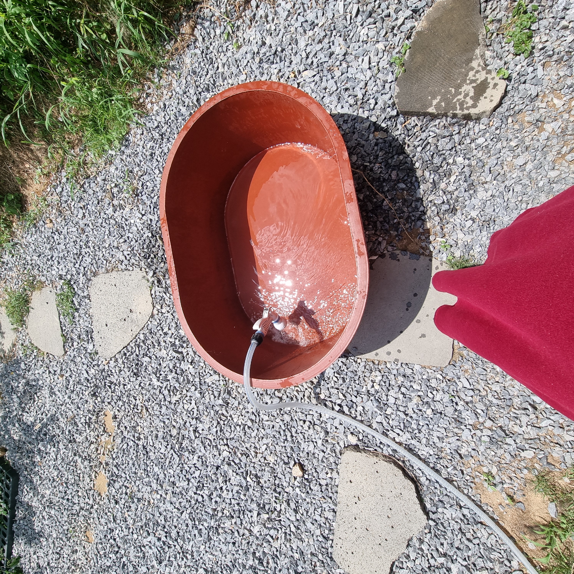 The image shows an outdoor scene on a sunny day. A large, oval-shaped, reddish-brown basin is filled with water. A white hose is feeding water into the basin, creating ripples on the surface. The basin rests on a concrete paver. The ground around the basin is covered with small grey gravel. Several irregular-shaped concrete pavers are scattered on the gravel, forming a path. To the right of the basin, a dense patch of green grass and other vegetation grows. In the foreground, a large portion of a dark red cloth, possibly a blanket or skirt, is visible. The scene appears to be in a rural or garden setting in Dunnae-myeon, South Korea, given the presence of gravel, stepping stones, and natural vegetation.