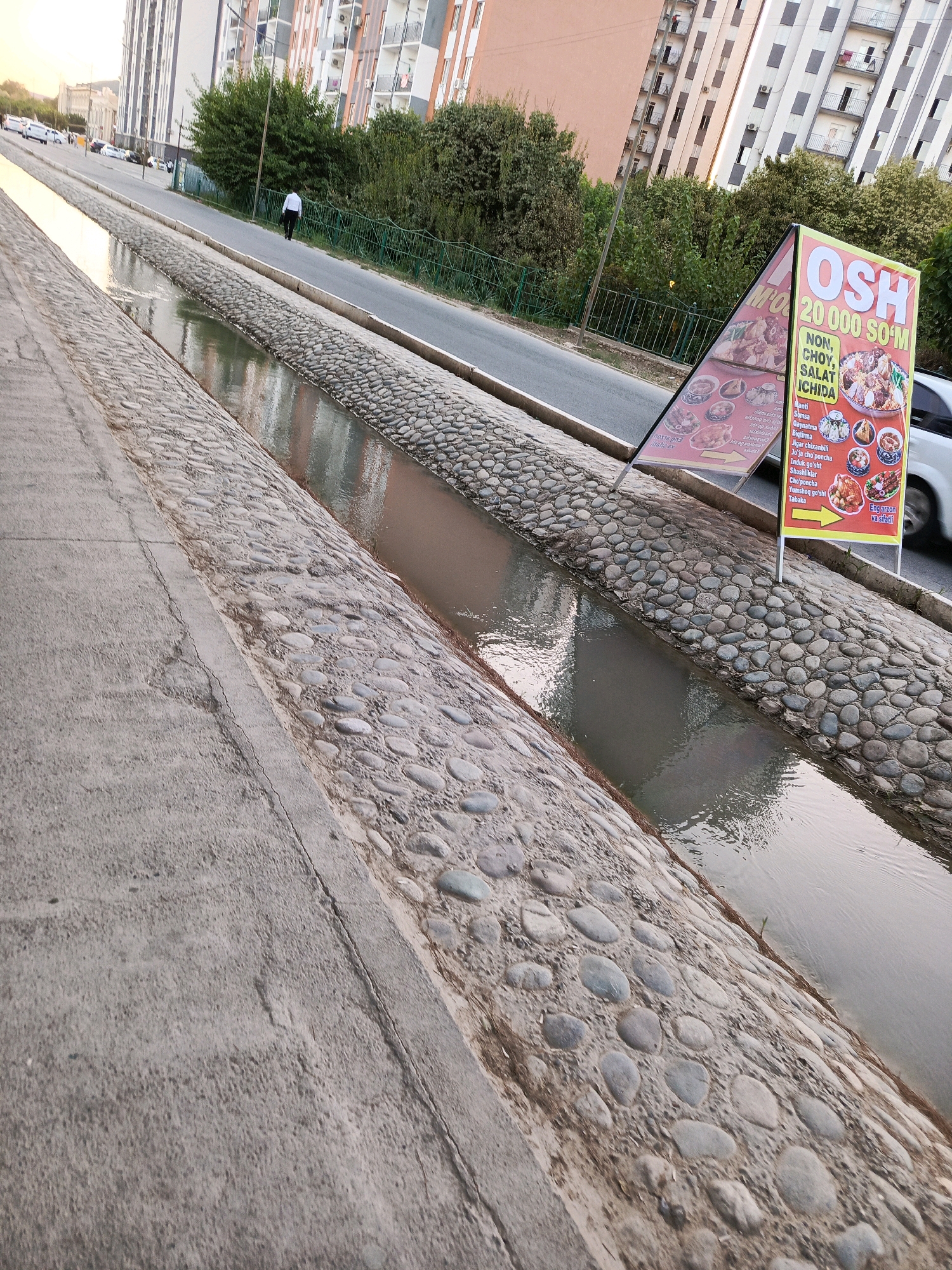 An outdoor, eye-level, medium shot shows a street scene next to a concrete and stone-lined drainage ditch with flowing water. In the middle distance, a person in a white shirt and black pants walks away from the camera on the paved sidewalk bordering the ditch. Buildings rise in the background, with trees and a green fence lining the street. To the right of the frame, a yellow A-frame sign advertises "OSH" with food images and text in an unknown language. A white car is partially visible to the right of the sign. The sky is overcast.